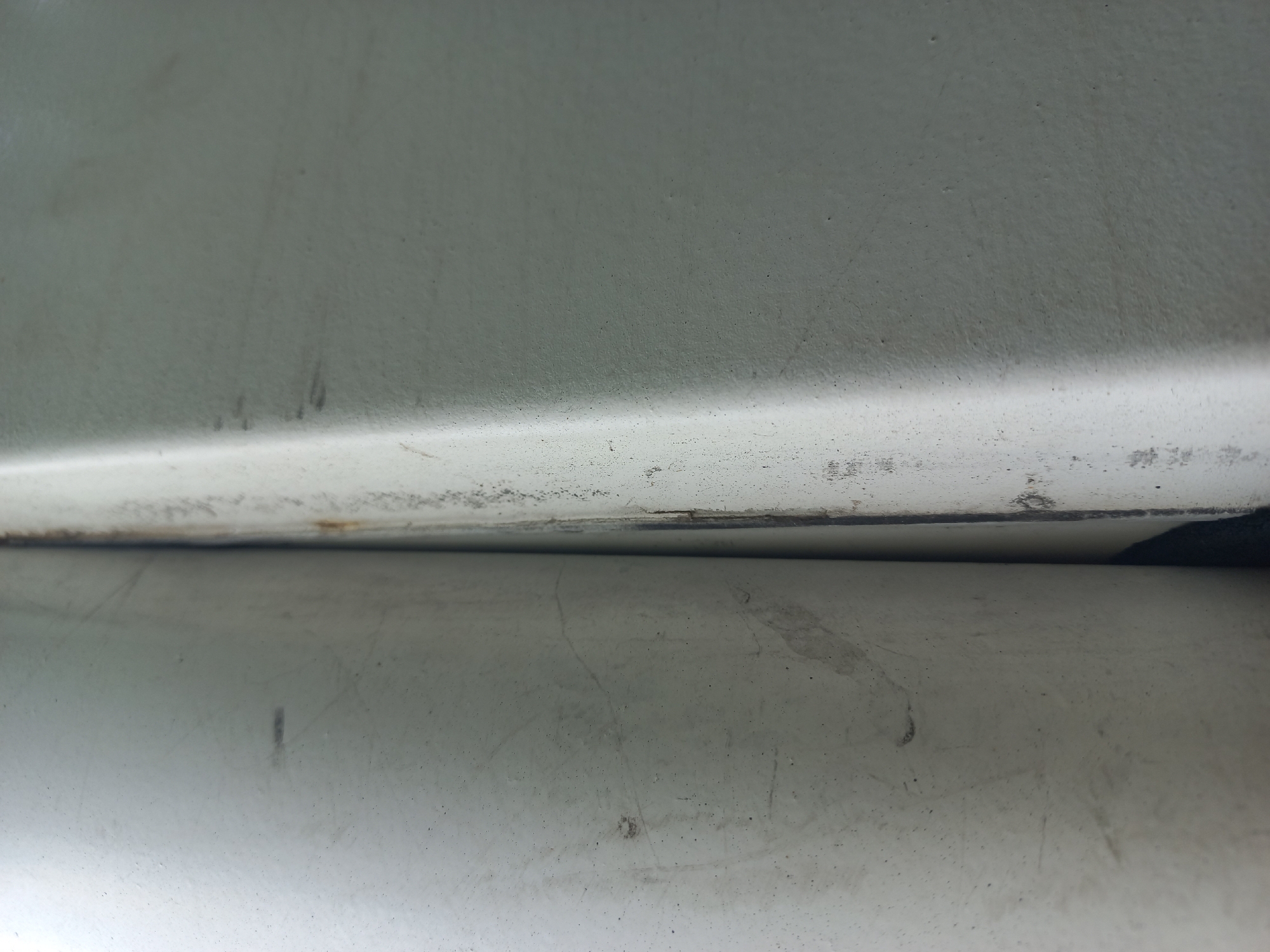 The image presents a close-up, abstract view of two adjacent surfaces, likely part of an exterior structure. The surfaces are painted a light, off-white color, exhibiting signs of wear and dirt. A thin, dark seam or gap separates the two surfaces, with some visible rust and discoloration along the edge of the right surface. The texture of the surfaces is slightly rough, with subtle undulations and small pockmarks. No people, animals, or specific landmarks are identifiable. The setting appears to be outdoors, given the natural lighting and the weathered appearance of the materials. The image lacks any context that would definitively place it within Velykodolynske, Ukraine.