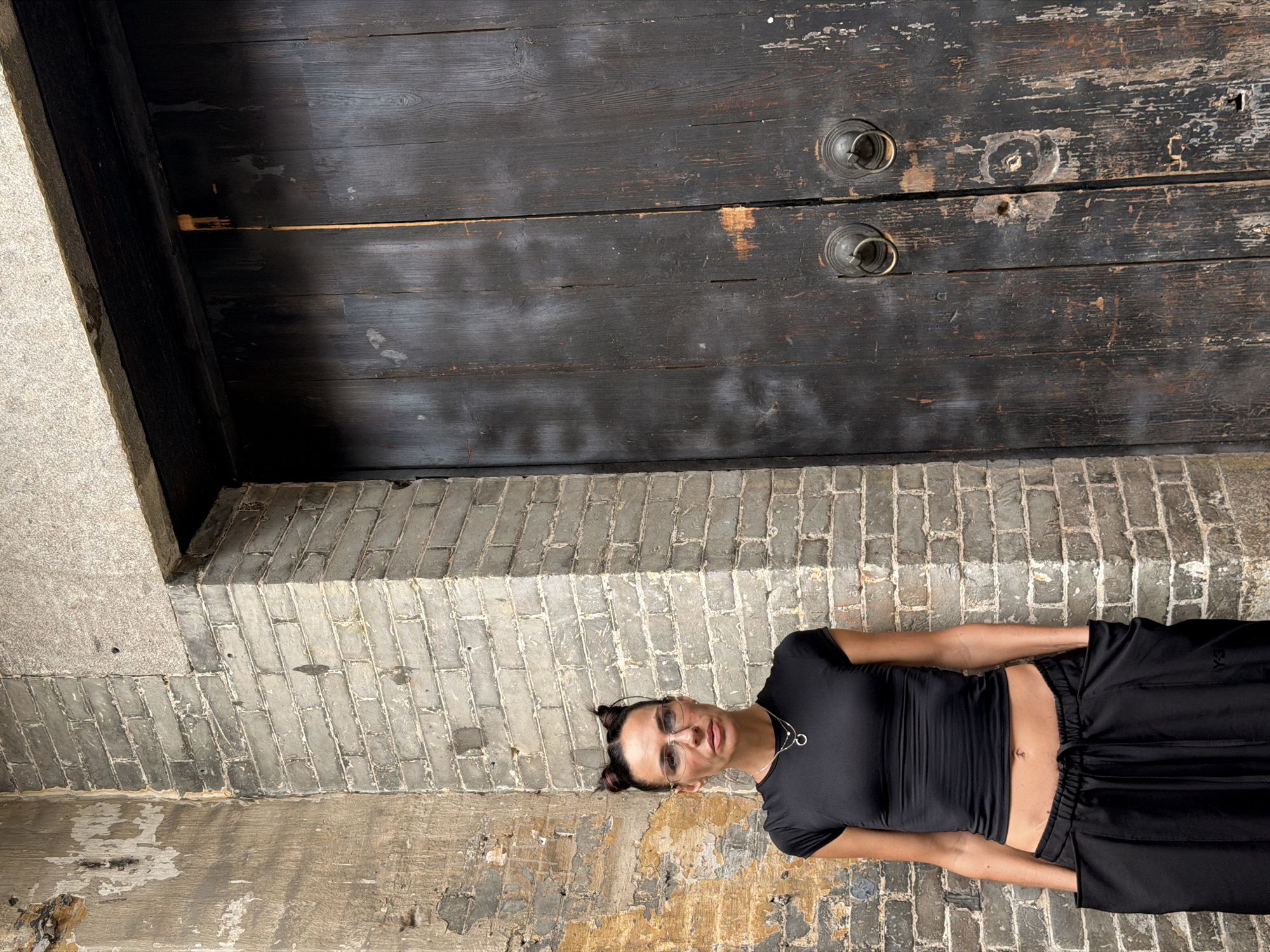 A woman stands against a brick wall in front of a large wooden door in Chancheng District, China. She is wearing a black crop top, black pants, and glasses. The door is dark wood with two round metal handles. The brick wall is grey and weathered. The scene appears to be in an outdoor urban environment.