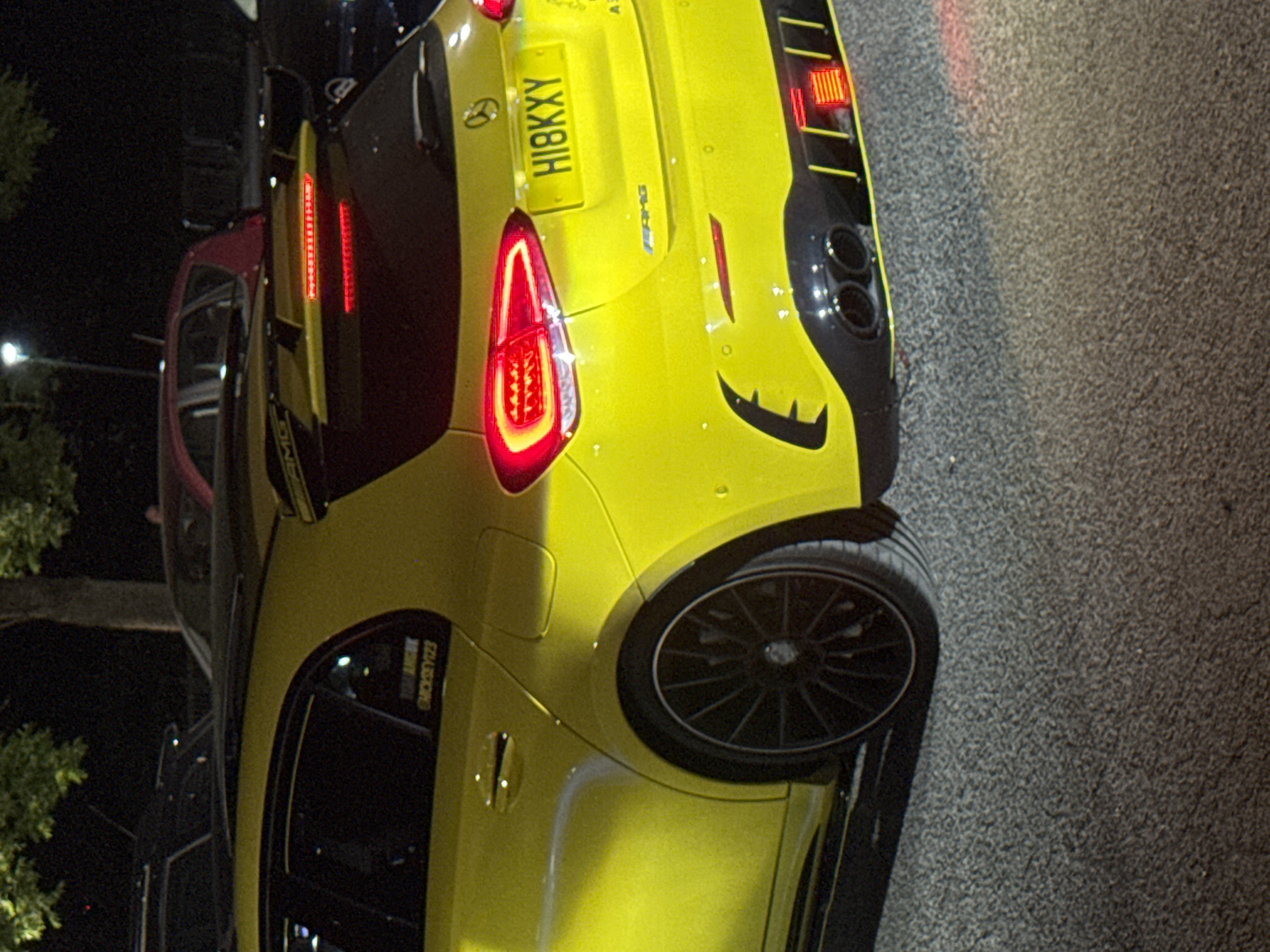 A yellow Mercedes-AMG A45 S car with registration number HI8KXY is parked on a street in the Borough of Spelthorne, United Kingdom. The rear taillights are illuminated. To the left is a car, visible only in part. The environment is urban with trees and street lights in the background. The surface on which the cars are parked is asphalt.