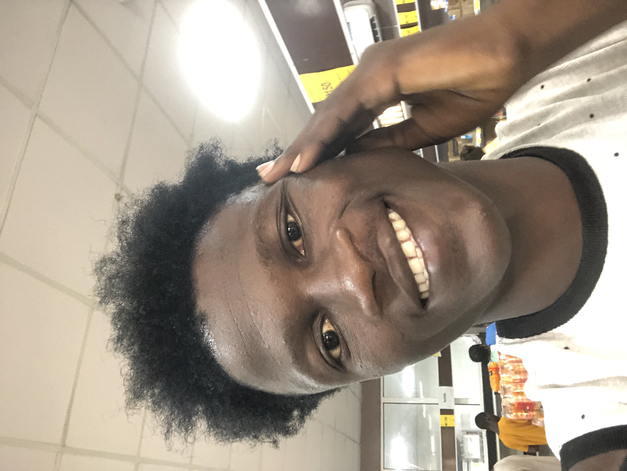 A young Black male with a large, bushy afro smiles into the camera, holding his right hand to his temple. He is wearing a white t-shirt with a black collar and small black polka dots. The background appears to be an indoor marketplace, with shelves of goods visible, including bottles of orange liquid. A yellow sign with "150" printed on it is visible on a shelf in the background, and an air conditioning unit is mounted on the wall. The lighting suggests it could be daytime, possibly from fluorescent ceiling lights. The image appears to be a selfie taken in Ado, Nigeria, based on the context provided.