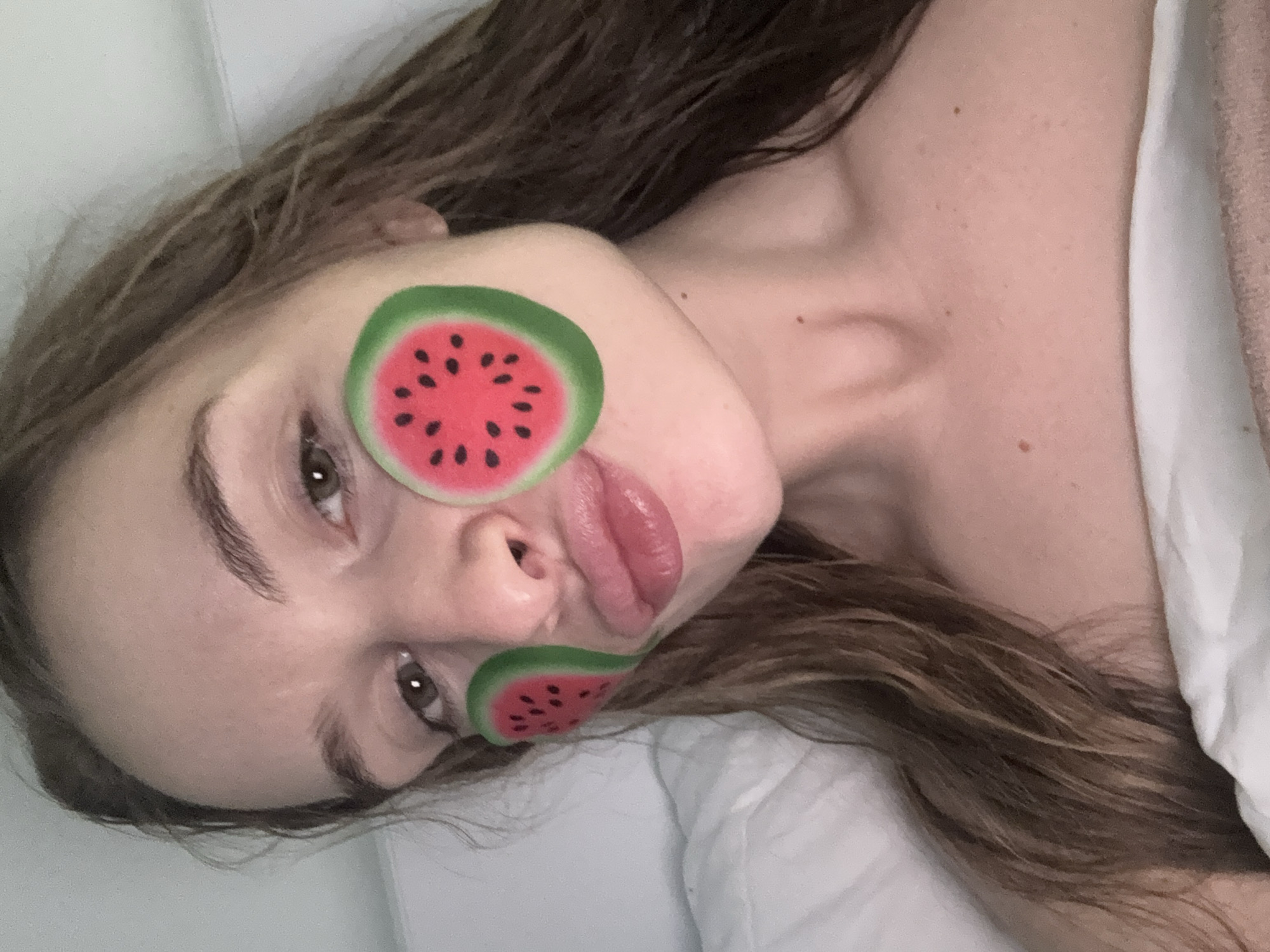 A young woman with long, brown hair is lying down with her head on a white pillow. She has two watermelon-shaped stickers on her face: one is positioned over her left eye, and the other is on her lower left cheek, near her mouth. The location is unknown; Budva, Montenegro, is not evident in the image.