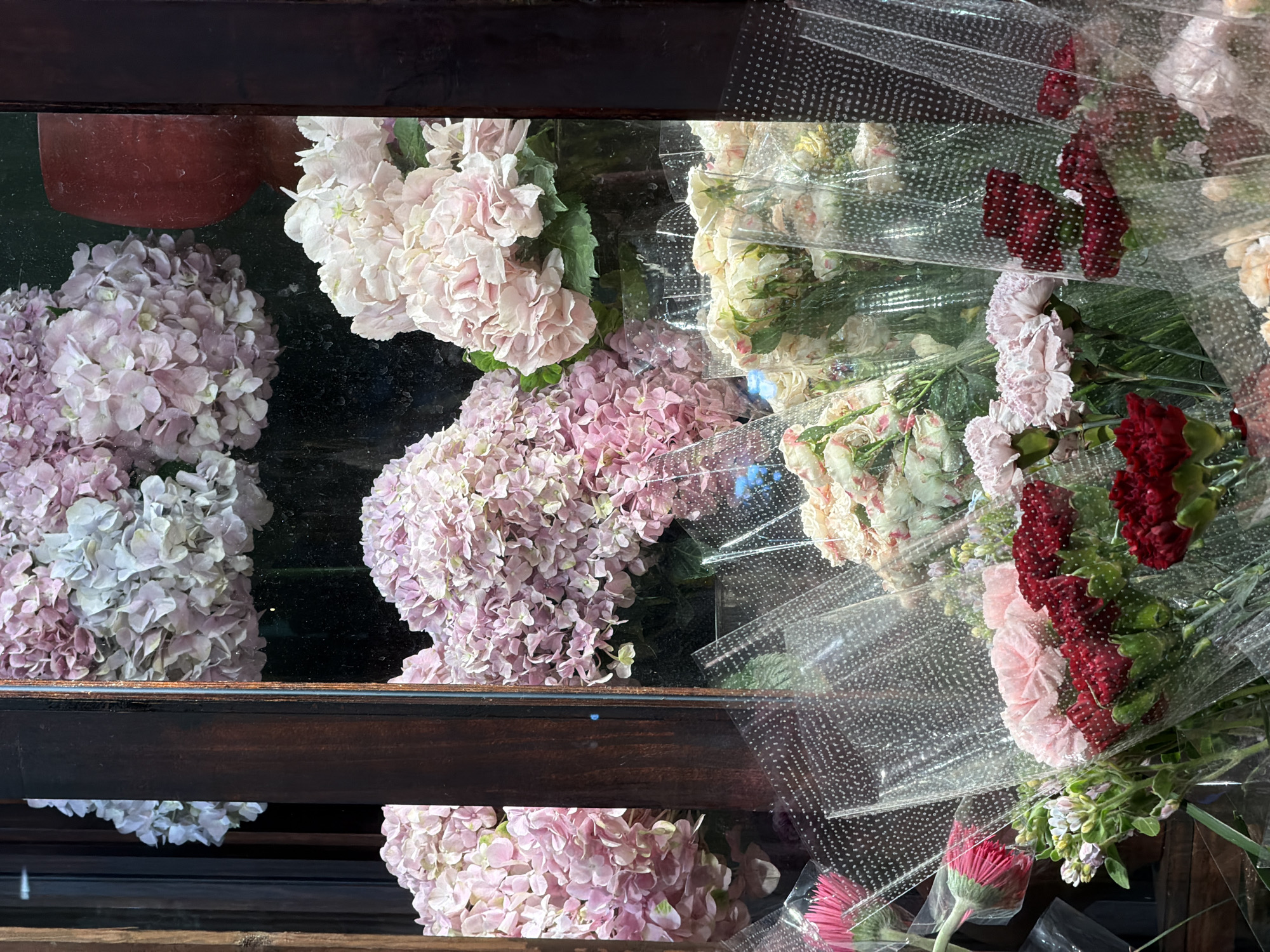 The image depicts a floral display, possibly in a flower shop. A variety of flowers including hydrangeas in shades of pink and white, as well as carnations and other unidentified flowers are arranged in what appears to be a tiered display case with a dark wooden frame. Some of the flower bouquets are wrapped in clear plastic. The location is Nha Trang Ward, Vietnam.
