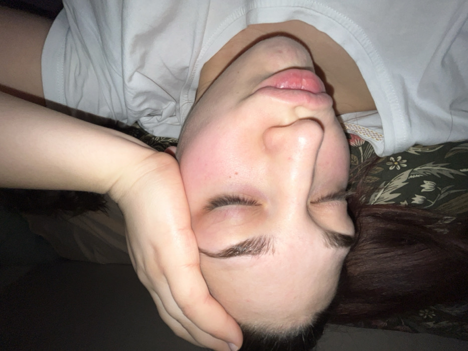 A person with closed eyes rests their face in their hand, lying down. They have fair skin, defined eyebrows, and slightly parted lips. A white t-shirt is visible at their neck. Part of their brown hair is visible on the right side. The background includes a bed with a patterned bedspread and a dark, undefined area. The perspective suggests the photo was taken from above.