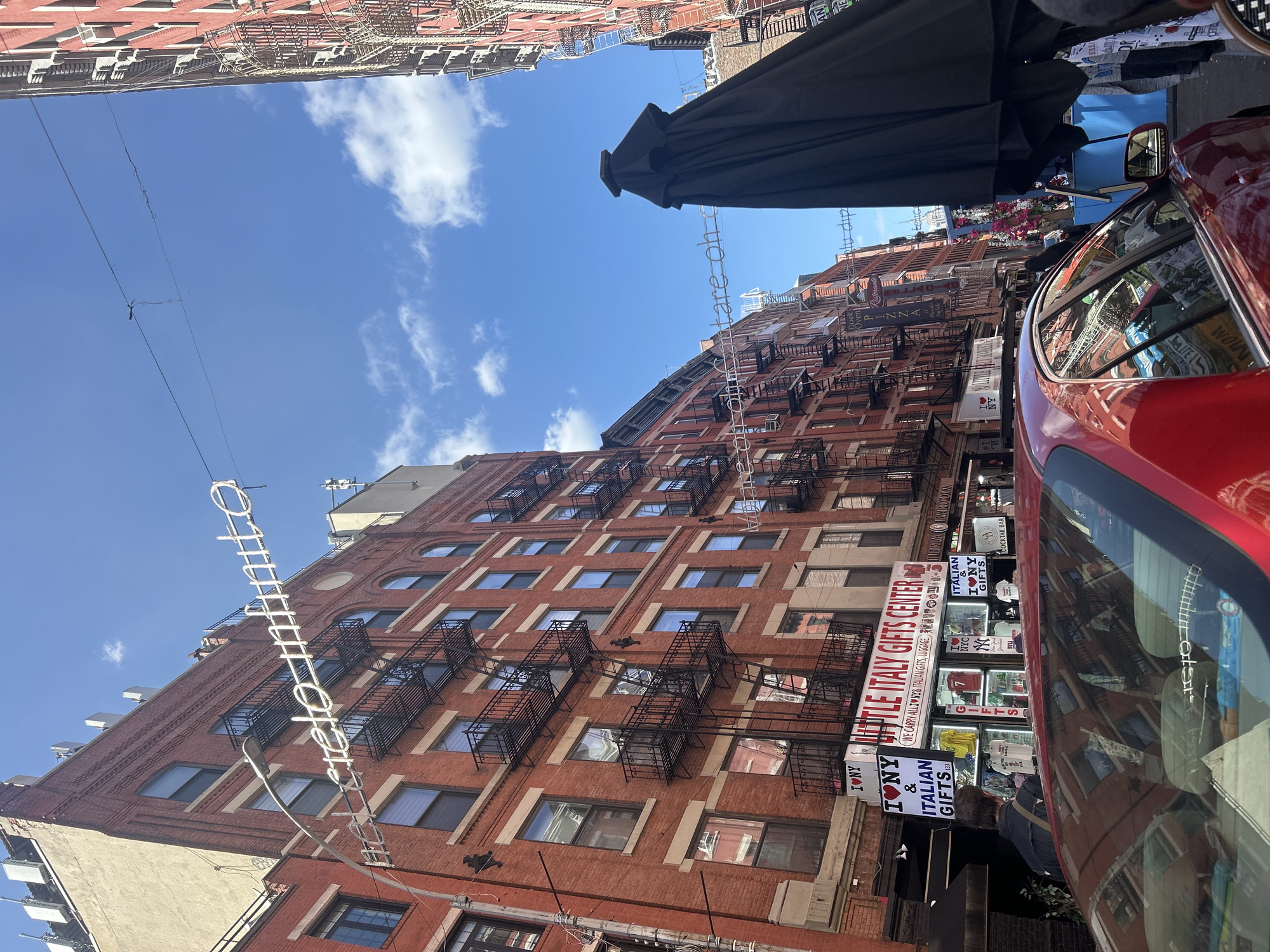 This image captures a street scene in the Little Italy section of New York City, United States of America. The perspective is from street level, looking up at the buildings, storefronts, and sky.
The focal point is a red-brick building adorned with multiple fire escapes on its facade. Signs for "Little Italy Gifts Center" and "Italian Gifts" are visible, indicating the commercial nature of the street. Adjacent to the building is a red vehicle, partially capturing the reflection of surrounding buildings and activity in its windows.
Above, a bright blue sky is dotted with a few clouds, providing contrast to the earth-toned buildings below. A black awning or umbrella partially obscures the top right corner of the frame. Banners and wires also stretch across the scene, contributing to the urban texture. Pedestrians can be seen browsing in the store, suggesting the usual activity within Little Italy.