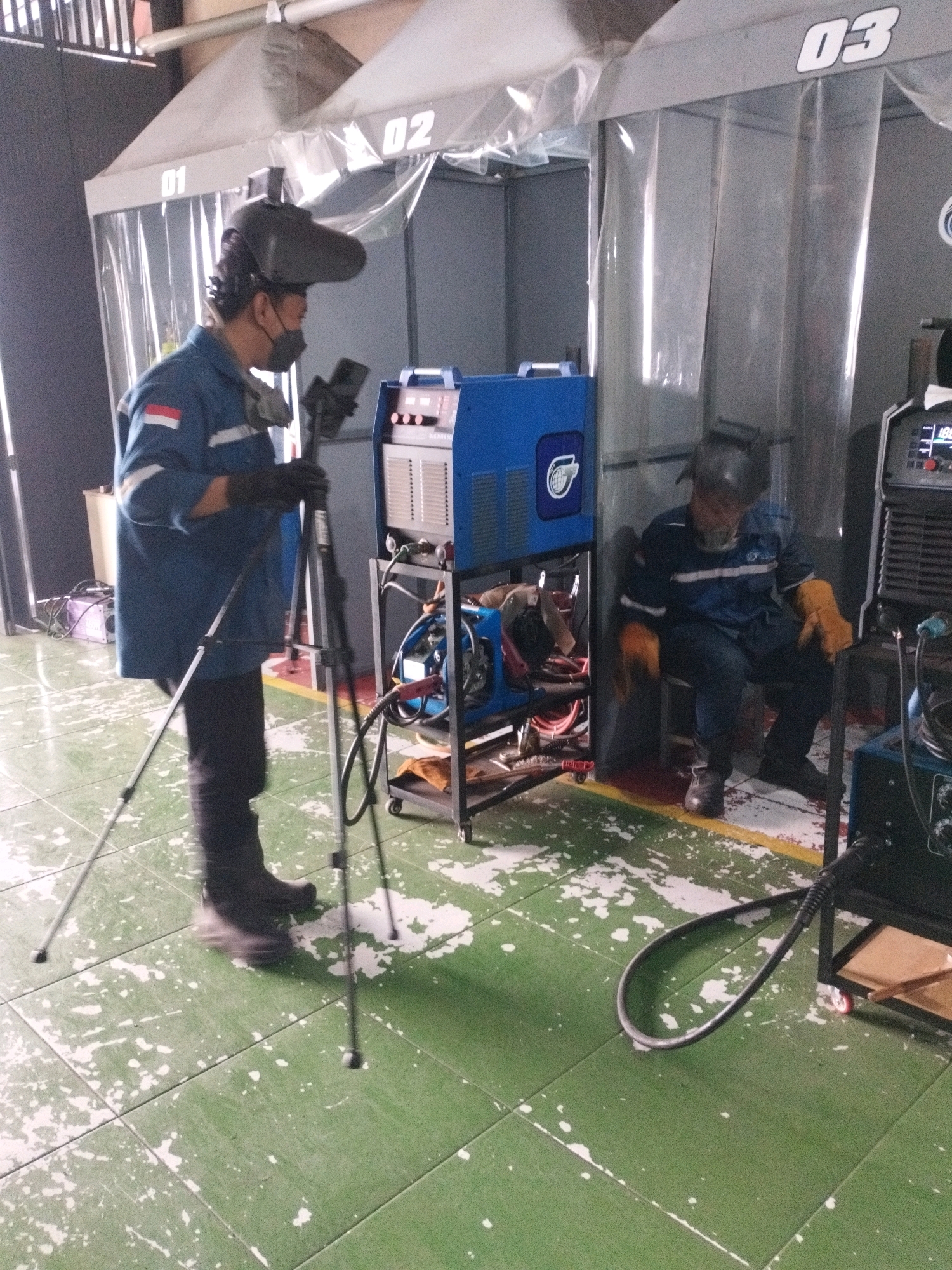 In Selomerto, Indonesia, within a vocational training facility, two individuals are engaged in welding activities. The scene is set in a workshop area delineated by numbered booths (01, 02, and 03 visible). A foreground figure, wearing a blue uniform with white reflective stripes and a welding helmet, is standing beside a tripod that holds a smartphone. They are holding welding gloves and appear to be setting up for or observing a welding task. To the right, another individual in a similar blue uniform and welding helmet is seated on a stool, also wearing orange welding gloves, with a welding machine in front of them. The environment features industrial-style flooring with green tiles and white speckled markings, suggesting a practical workshop setting. Welding equipment, including welding machines and cables, are visible throughout the space, indicating active use. The lighting appears to be overhead industrial lighting.
