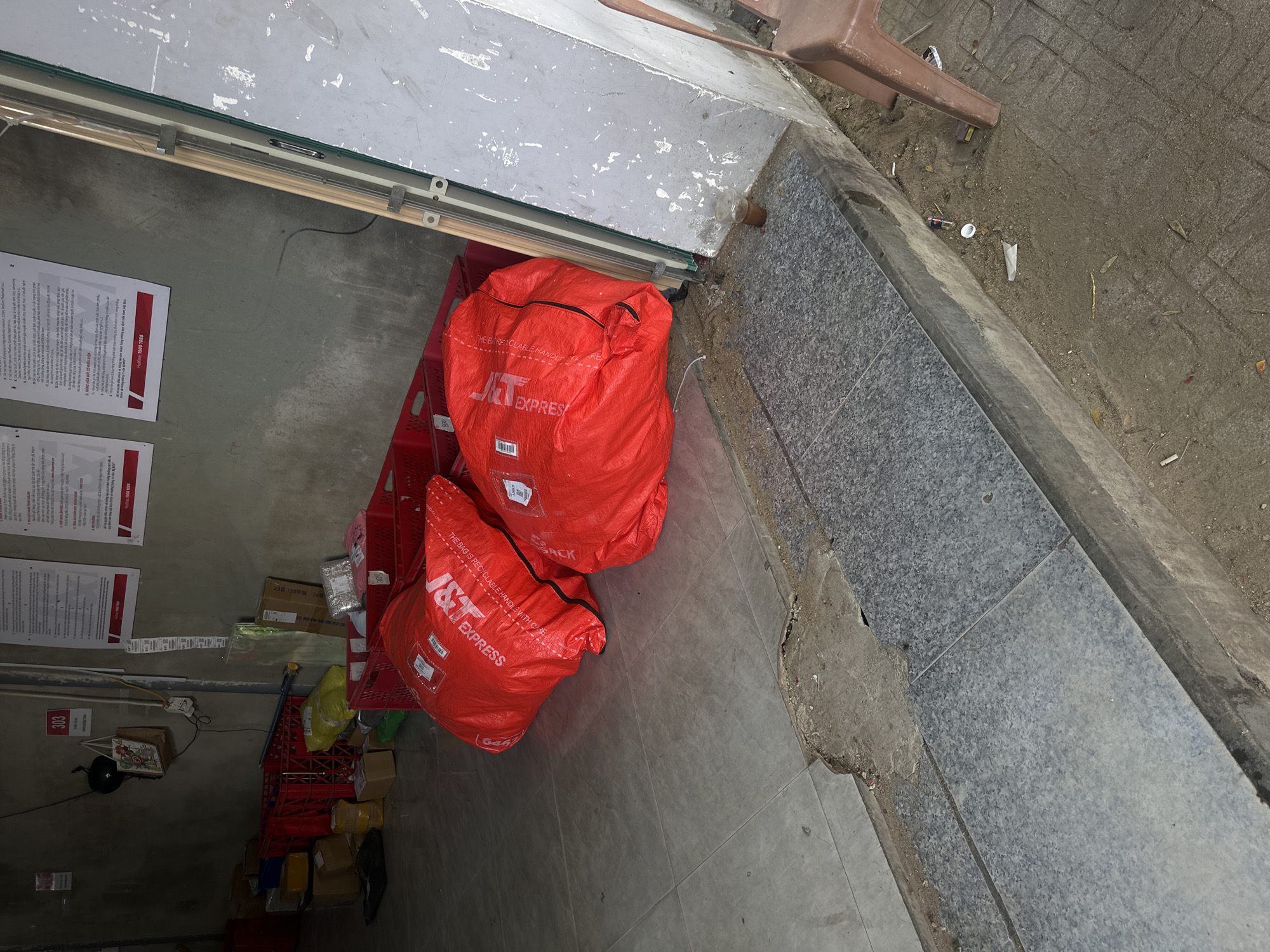 The image depicts an outdoor scene, likely a storefront or loading area, in Bac Nha Trang Ward, Vietnam. Two large, bright red J&T Express packages are prominently displayed on the tiled floor in the foreground, suggesting a delivery or logistics hub. The packages are made of a durable, possibly synthetic material, and are sealed with zippers and labels. Behind the packages, several red plastic crates are stacked, some containing yellow and other colored items, indicating storage of goods. In the background, a concrete wall displays several white posters with text and graphics, likely official notices or advertisements. The overall environment appears utilitarian, with a mix of tiled and concrete surfaces, and evidence of daily operations, such as the presence of shipping materials.