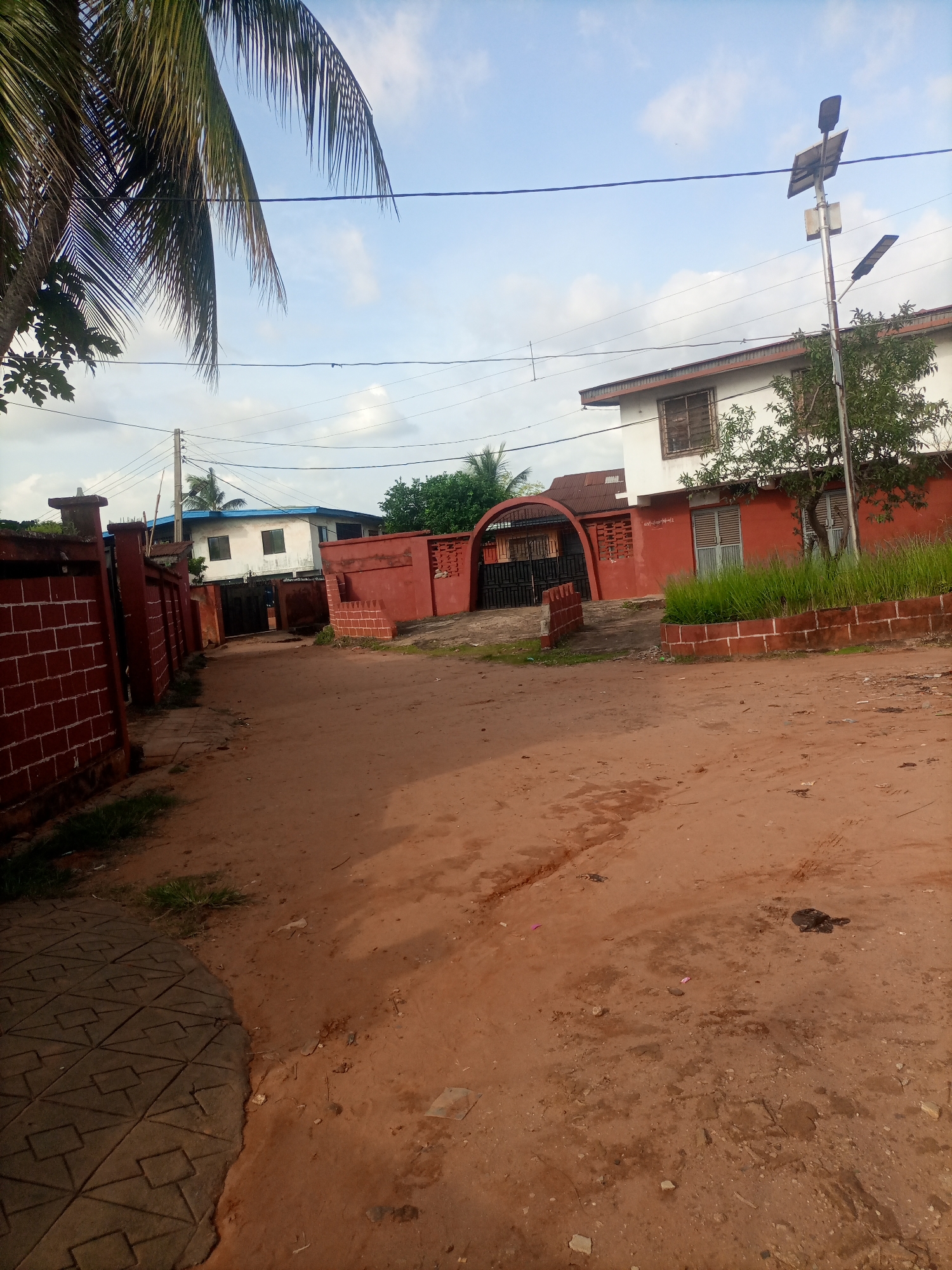 This image depicts a street scene in Nkpor, Nigeria. The foreground is dominated by a reddish-brown dirt road, with a section of patterned paving stones on the left side. In the background, there are several buildings. On the left, a red brick wall with pillars lines the street. Further down, a white building with a blue roof is visible behind a red gate. To the right, a two-story building with white and red walls features barred windows and a red tiled roof. A solar-powered street light stands prominently in front of this building. Lush green grass grows in patches along a low red brick wall in front of the building. A palm tree extends from the left edge of the frame, with its fronds reaching towards the sky. The sky is a pale blue with scattered white clouds, suggesting it is daytime, likely late afternoon given the lighting. There is no visible activity or people in the image. The overall impression is a quiet residential or commercial area. No text is visible in the image.