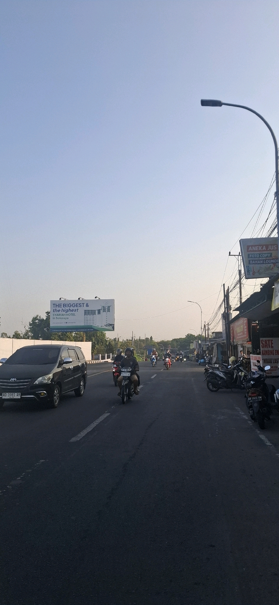 The image depicts a street scene in Colomadu, Indonesia, likely during the late afternoon or early morning given the lighting. A black Toyota Innova car with license plate AD 1981 AM is stopped on the left side of the road. Several motorcycles are present, with many riders wearing helmets. Some motorcycles are in motion on the road, while others are parked on the right side. On the left side of the road, a billboard advertises "THE BIGGEST & the highest SYARIAH HOTEL In Soloraya". On the right side of the road, a sign for "ANEKA JUS FOTO COPY BAHAN LAUNDRY" is visible, along with other businesses and signs, including one that reads "SATE KAMBING". The sky is clear and a light blue. A street lamp is visible on the right side of the frame. The road surface appears to be asphalt and shows signs of traffic, including tire marks and scattered debris. Trees are visible in the background on the left.