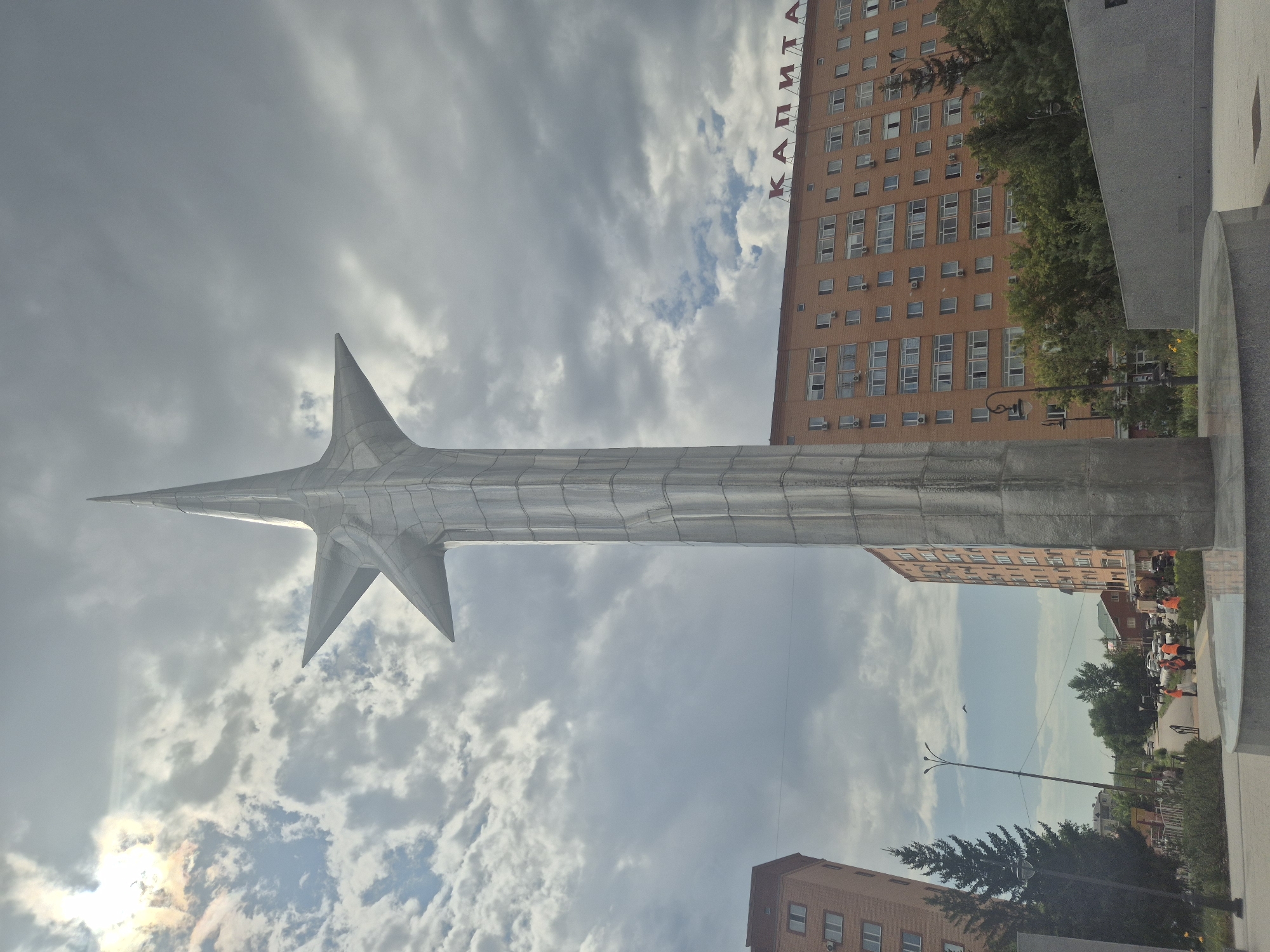 The image captures a monument in Kokshetau, Kazakhstan, a large, metallic, star-shaped sculpture reaching towards a cloudy sky. The sculpture is composed of many facets, giving it a geometric and modern appearance. In the background, there are two multi-story brick buildings, one on the left and a larger one on the right. The building on the right has the word "КАПИТА" (Kapita) written in red letters across its top. Trees and streetlights are visible between the buildings and in the foreground. Several people wearing orange vests are present near the base of the sculpture, possibly working on or around it. The overall scene depicts an urban environment with a prominent public art installation.
