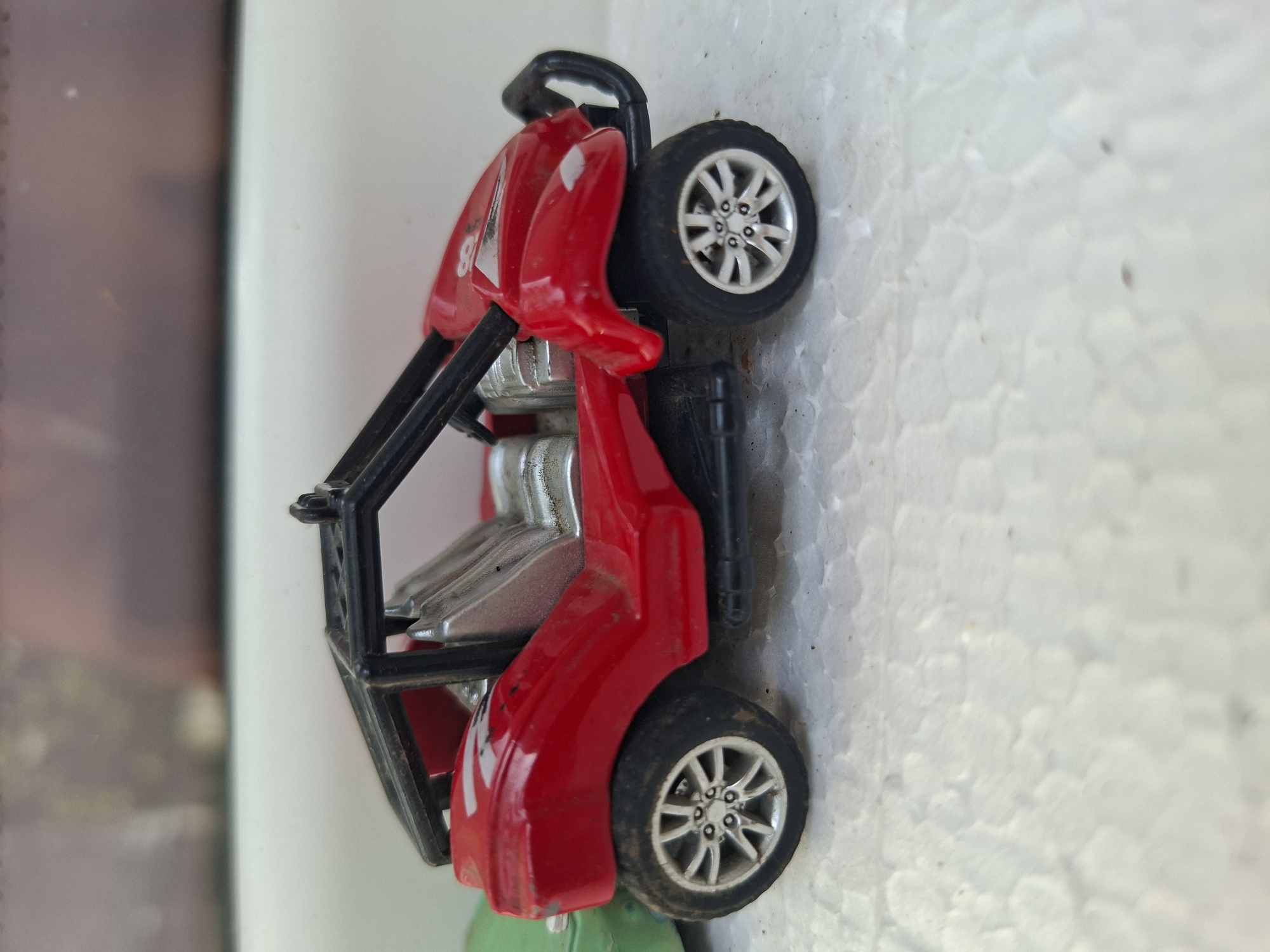 A red toy dune buggy is positioned on a white textured surface. The dune buggy has a black roll cage, silver seats, and silver rims on its tires. A portion of a green toy is visible to the left of the dune buggy. The background consists of a blurry surface with muted colors. The location in Gorkovskiy, Russia, is not discernible from the image.