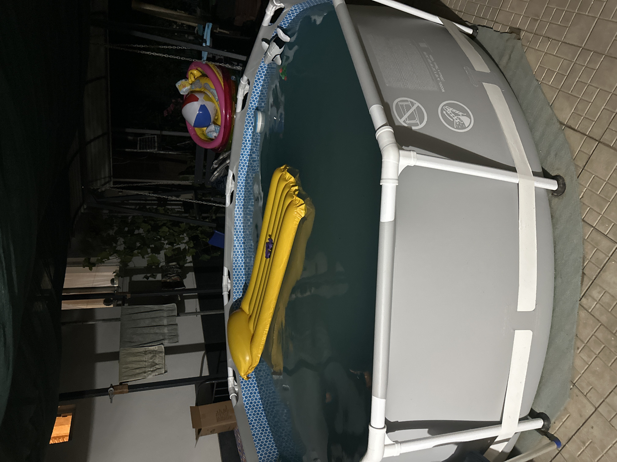 This media file shows an above-ground swimming pool set up outdoors at night. The pool has a grey exterior and a blue and white patterned interior liner. A yellow inflatable pool float is partially submerged in the water. To the left of the pool, a beach ball and an inflatable ring are suspended by chains, possibly as decorations or for play. In the background, there is some foliage and what appears to be a metal structure, perhaps a canopy or shelter. A cardboard box is visible on the ground near the pool. The scene is illuminated by artificial light, indicating it is nighttime. The ground surrounding the pool is paved with square tiles. The location is Pîrlița, Moldova.