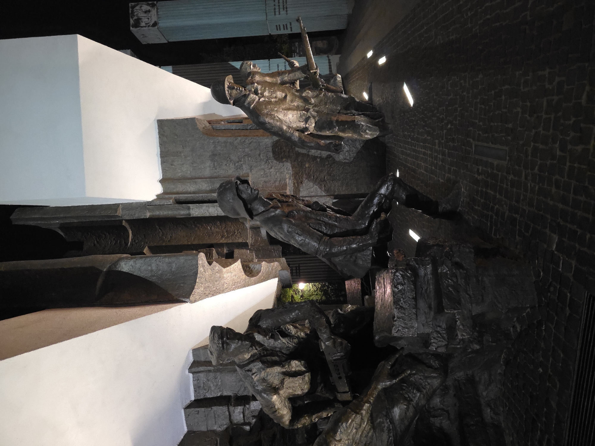 The image depicts a sculptural monument in Warsaw, Poland. The monument features three figures, likely soldiers, in varying states of distress or death. One figure lies prone, another is seated with their head bowed, and the third stands with a rifle. The sculptures are dark in color, possibly bronze or a similar metal, set against a rough, stone-like background with small recessed lights. Part of the monument includes a white, geometric structure resembling steps or platforms. The overall setting is dark and somber, appropriate for a memorial. The presence of brickwork suggests an urban, possibly historical, location.