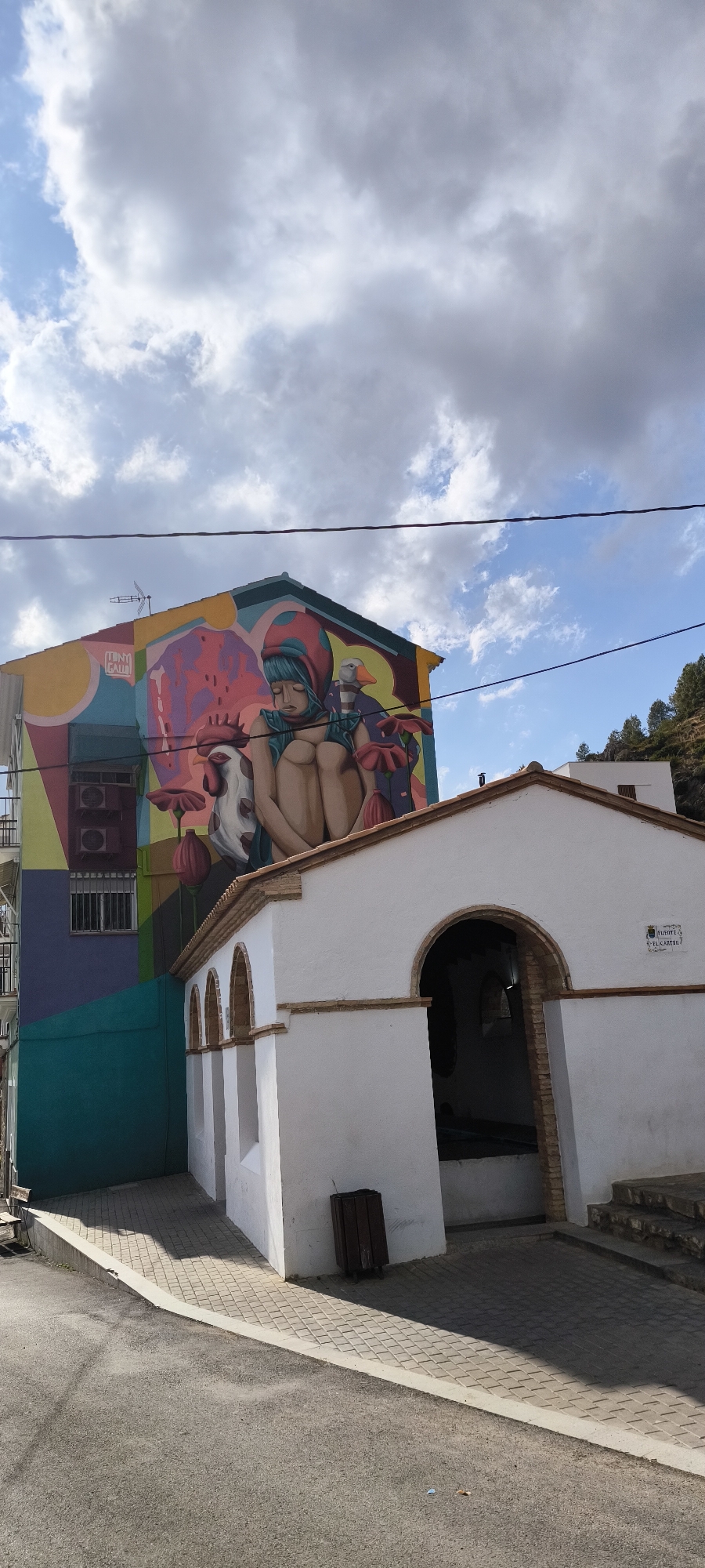 The image shows a vibrant mural painted on the side of a building in Fanzara, Spain. The mural depicts a figure, possibly a child, with blue hair, wearing a red hood, and sitting in a fetal position. The figure is surrounded by stylized floral elements and other characters, including a rooster and a duck. The building on which the mural is painted is colorful and geometrically patterned. Adjacent to this building is a smaller, white structure with arched openings, possibly a public fountain or shelter, indicated by a sign that reads "FUENTE EL CARTAN." The scene is outdoors, with a blue sky dotted with clouds visible in the background, and trees on a hillside in the distance. A paved pathway leads to the white structure, and a road is in the foreground. A dark-colored trash receptacle is placed near the entrance of the white structure. Electrical wires are visible strung across the top of the frame.