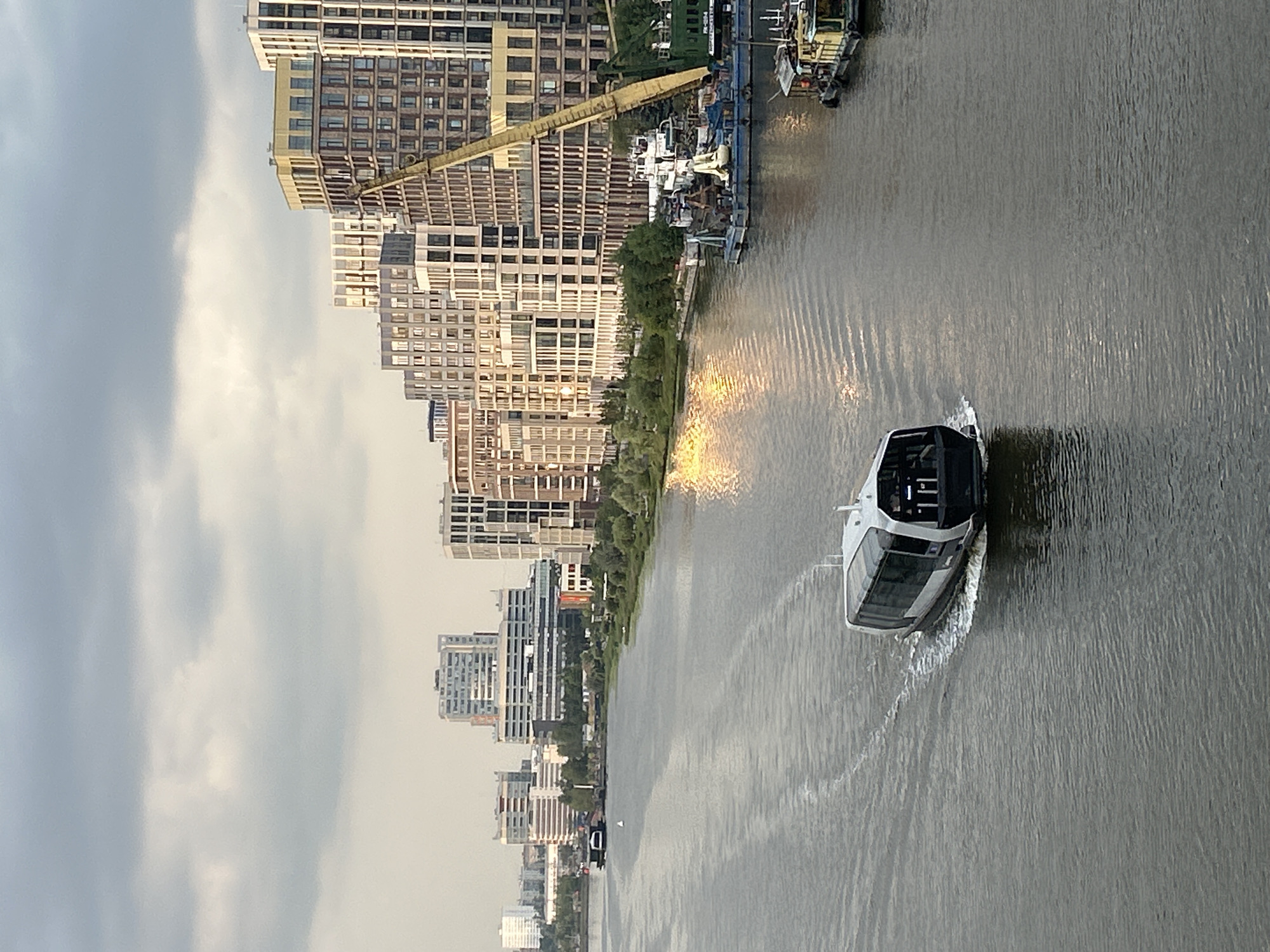 A contemporary, dark-hulled watercraft with a white, glass-enclosed cabin navigates a wide, calm river, leaving a white wake behind it. The vessel is positioned in the left foreground, moving towards the right and away from the viewer. The river's surface reflects the overcast sky and a bright, shimmering patch of sunlight. Along the right bank of the river, a dense line of modern, multi-story buildings with varied facades and window arrangements creates an urban backdrop. A tall, yellow construction crane is prominently visible amidst the buildings. Further to the left, across the river, a more distant urban skyline features several tall, rectangular buildings with numerous windows. The overall environment suggests a city on a waterway under a cloudy sky.