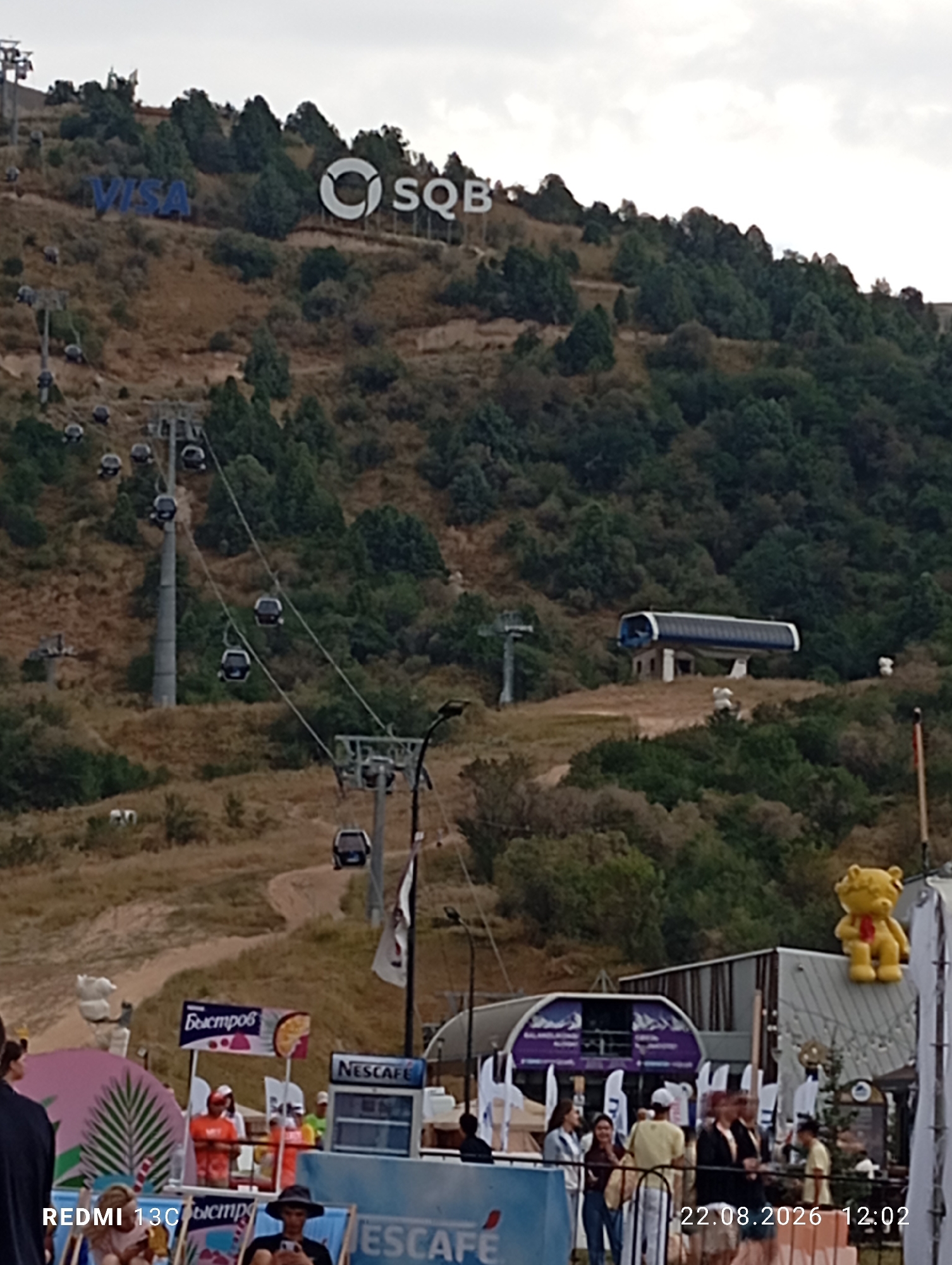 A wide shot captures a ski resort in Amirsay, Uzbekistan. A cable car system traverses a grassy, tree-dotted mountainside, with several cabins suspended mid-air. At the summit, the logos "VISA" and "SQB" are visible against the sky. A modern-looking cable car station is situated on the slope, and a yellow teddy bear figure with a red scarf is perched atop a building in the foreground. In the lower portion of the image, people are gathered near structures displaying various signs, including "Nescafe" and Cyrillic text. The scene is dated "22.08.2026 12:02" and tagged "REDMI 13C." The overall environment suggests an active outdoor recreational area during daytime.