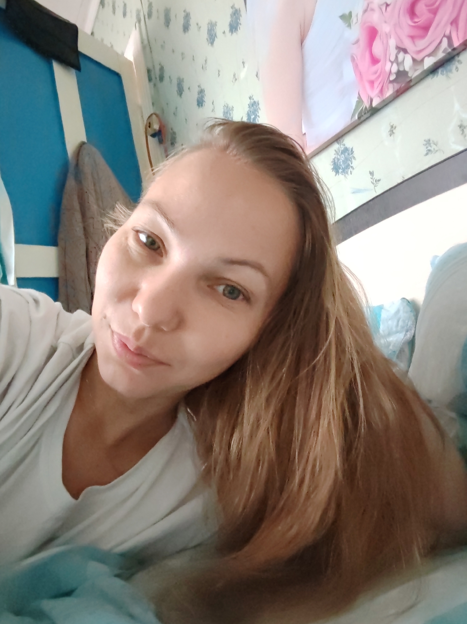 A young woman with blonde hair and green eyes is lying down, her head tilted to the side. She is wearing a white t-shirt and is partially covered by a light blue blanket. The background appears to be an indoor setting with a blue wall and floral wallpaper. The lighting suggests it is daytime. There are no discernible urban elements to indicate a specific location within the city of Chukhloma, Russia.