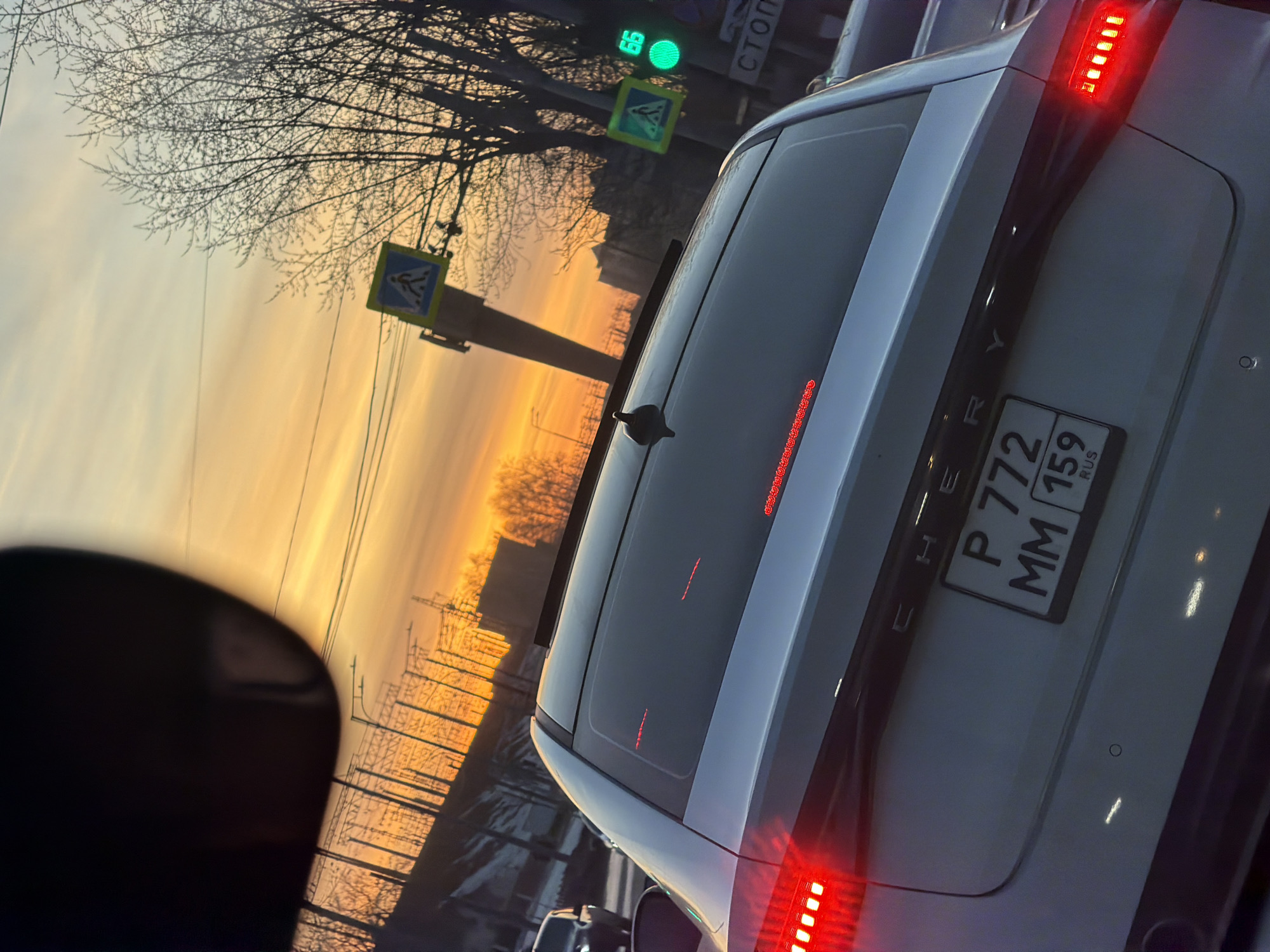 The image shows the rear of a white Chery car with license plate P772MM159 RUS. It is daytime, and the sun is setting or rising, casting an orange and yellow glow across the sky. A pedestrian crossing sign is visible in the background, along with bare trees and power lines. A traffic light shows a green signal. The location is inferred to be Perm, Russia based on the license plate information. The image appears to have been taken from inside another vehicle.