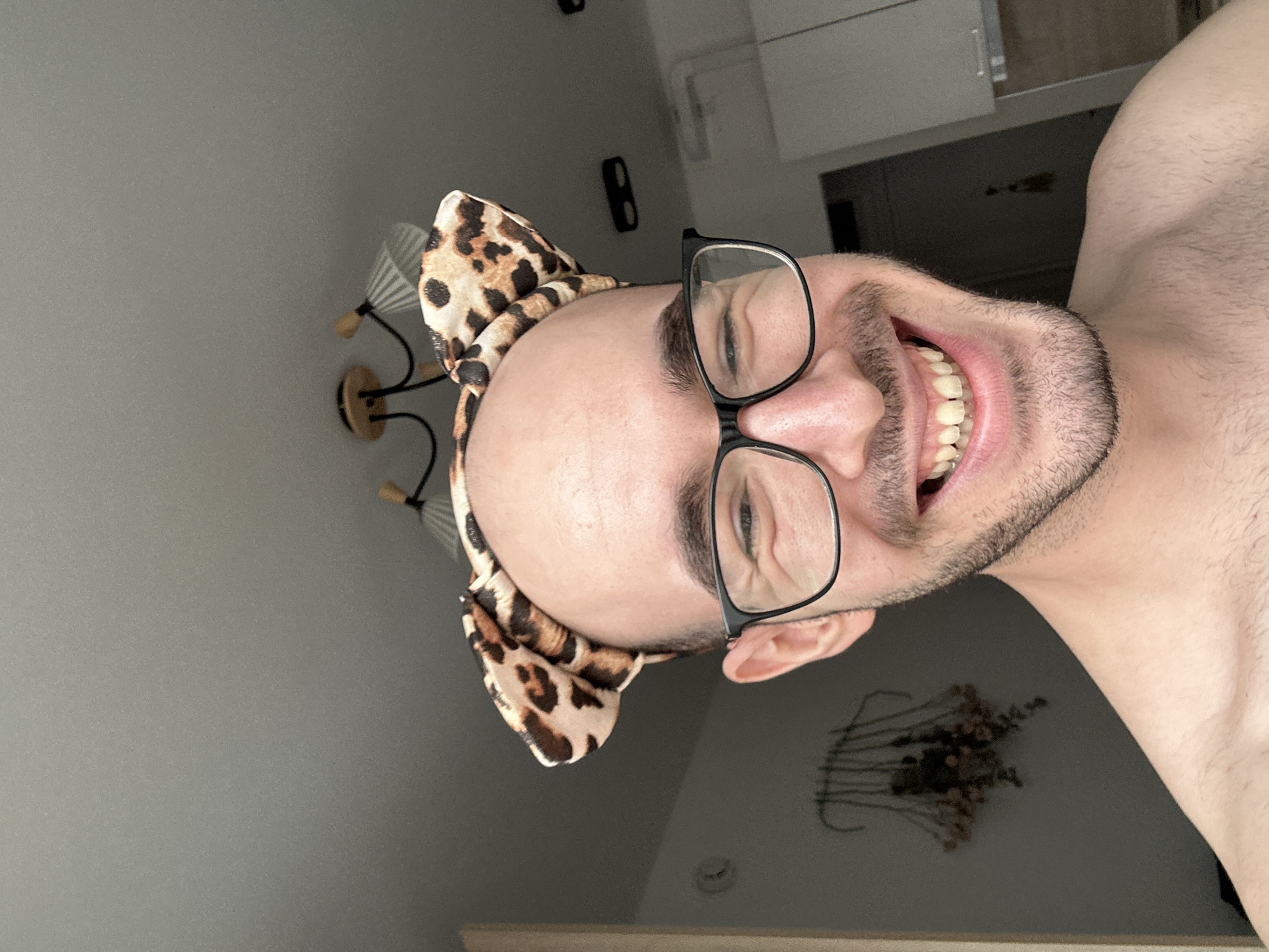 A selfie-style photograph depicts a shirtless person with light skin, wearing glasses and a leopard-print headband with bow-shaped details. The individual has a receding hairline, dark eyebrows, and a short, dark beard. They are smiling, revealing their teeth. The background includes a light gray ceiling and wall, a wall-mounted light fixture with multiple shades, a door, and a small wall decoration that appears to be a decorative dried flower arrangement. The location in the city of Lyubertsy, Russia is not discernible from the image.