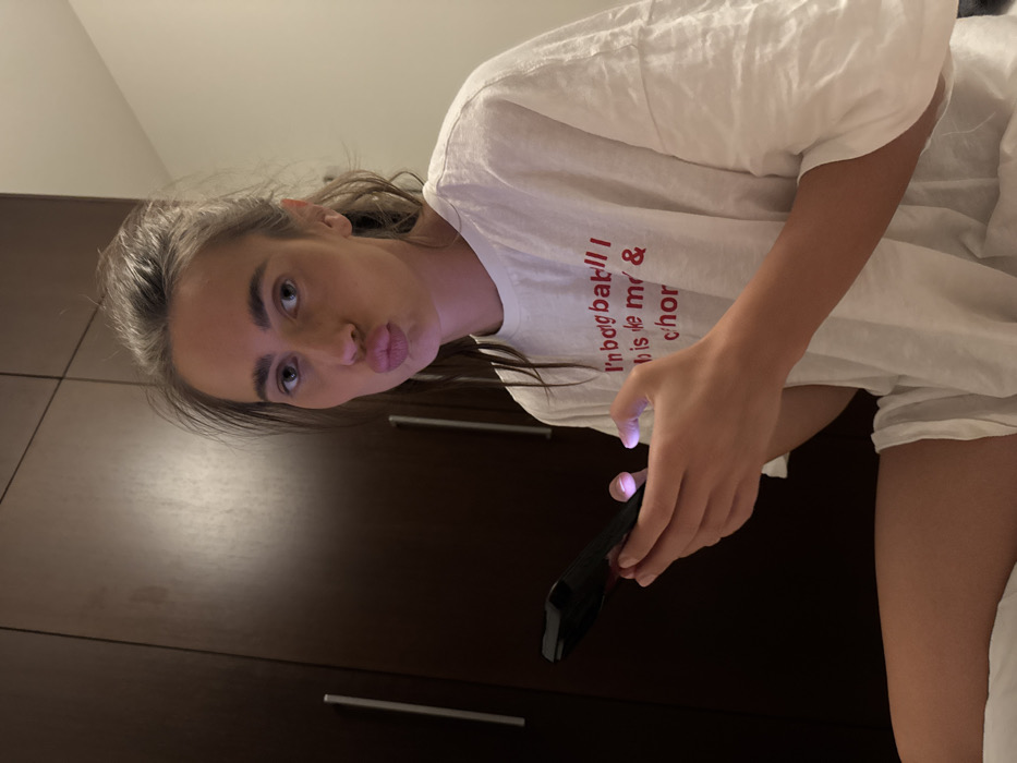 A young woman with light brown hair and blue eyes poses in a white t-shirt with red text. She’s holding a black smartphone, illuminated by the screen's glow. Her lips are pursed in a kissing face. Behind her is a dark brown wooden cabinet with sleek silver handles. The overall lighting is soft and warm.