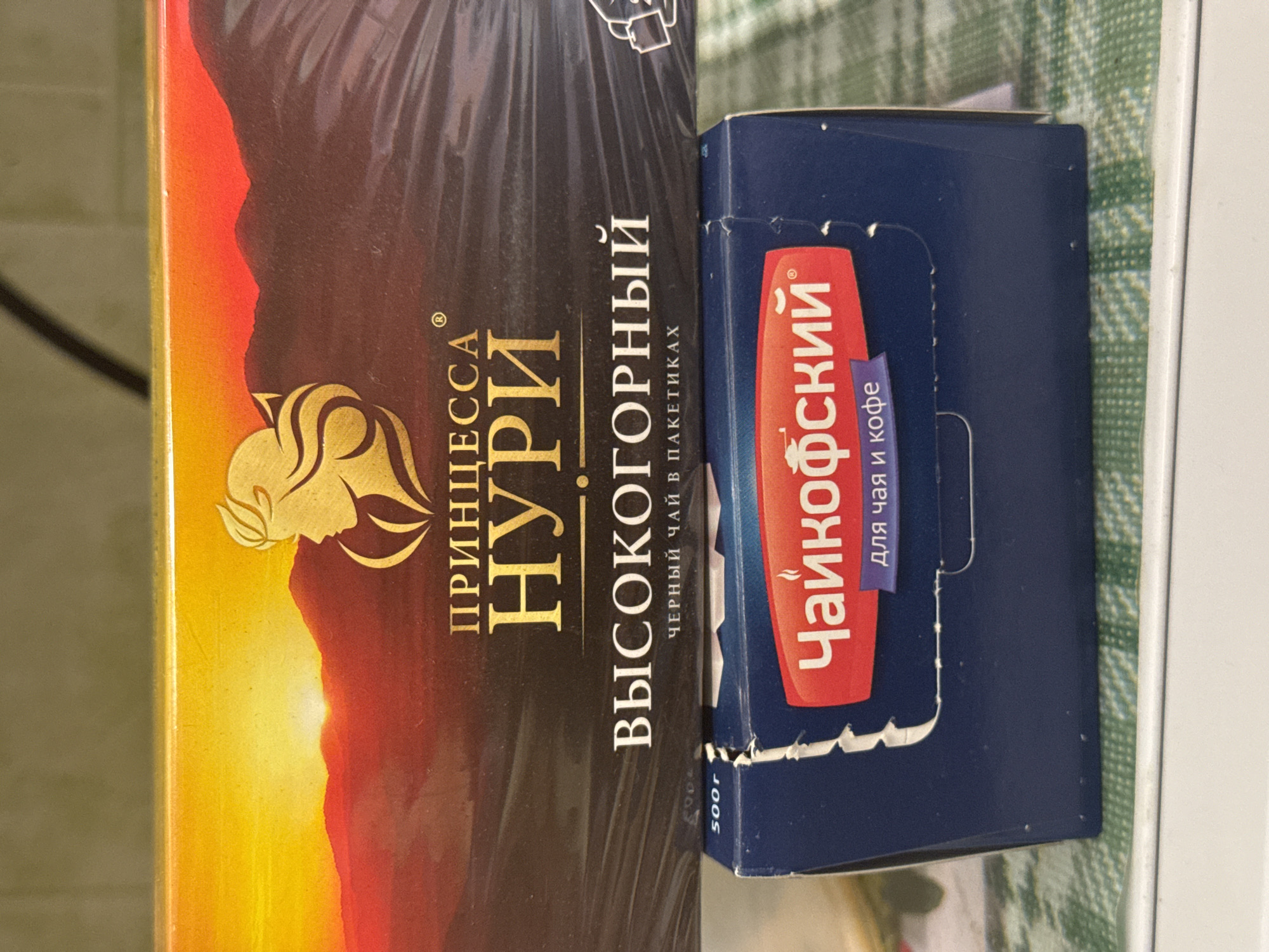 The image shows two boxes of tea. The box on the left is taller and features a graphic of a woman's profile against a sunset background. The brand name "ПРИНЦЕССА НУРИ" (Princess Nuri) and the words "ВЫСОКОГОРНЫЙ" (High Mountain) and "ЧЕРНЫЙ ЧАЙ В ПАКЕТИКАХ" (Black Tea in Bags) are written in Russian. The box on the right is blue and features the brand name "Чайкофский" (Tchaikovsky) in red, along with the phrase "для чая и кофе" (for tea and coffee) in Russian. The text "500 г" (500 g) can be partially seen on the boxes. Based on the brands of tea and the Cyrillic text, this image is likely taken in Russia. There is no evidence of a specific location in Chuprino.