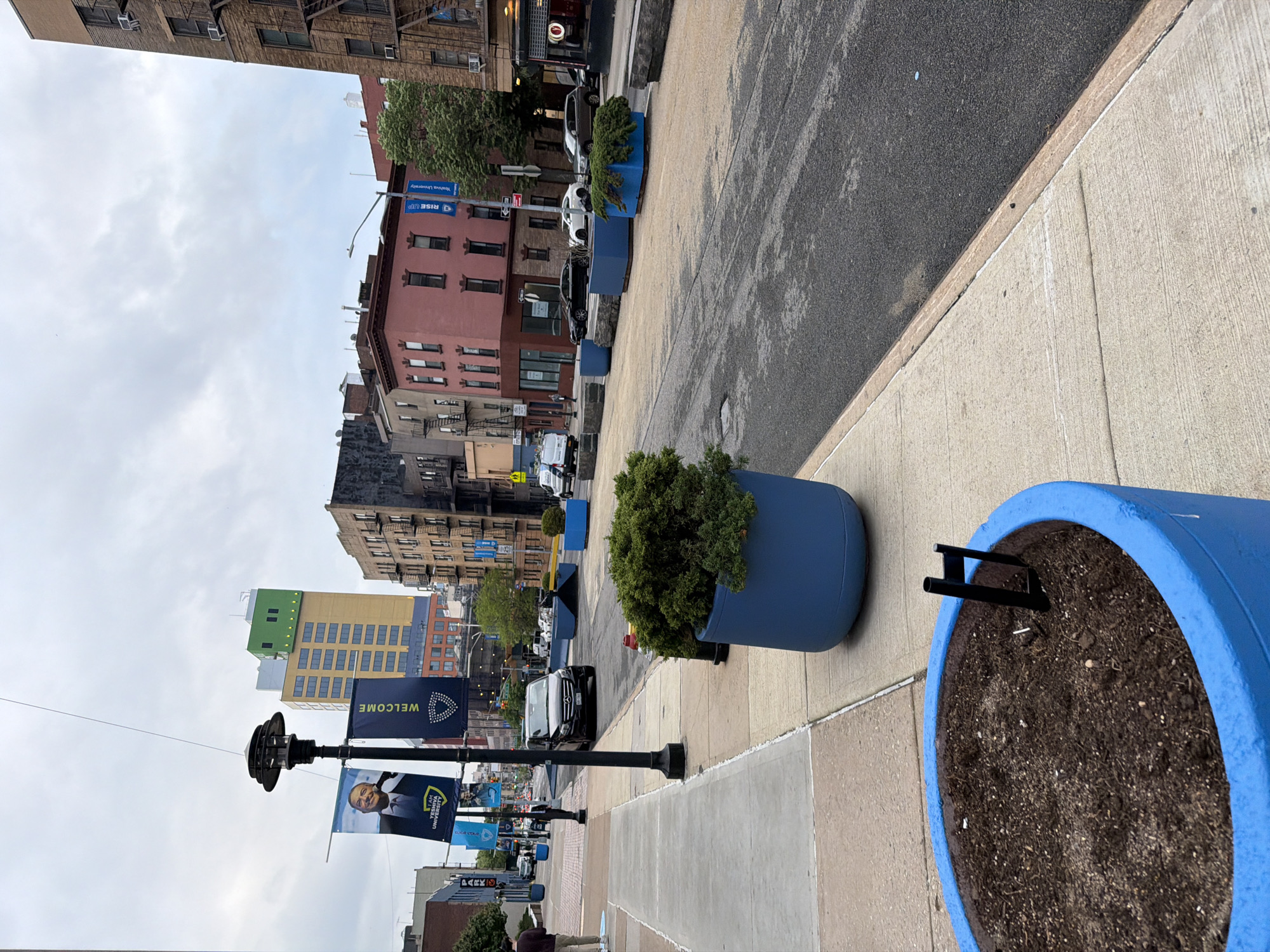 The image captures an urban streetscape in New York City. The foreground features large, bright blue planters, some filled with soil and others with shrubbery. These planters line the edge of a wide sidewalk made of concrete slabs. Behind the planters, a grey asphalt street stretches into the distance, bordered by buildings of varying heights and architectural styles. Buildings are brick and stone with different colored finishes. One yellow building has a green facade. Street signs are visible. Flags or banners with "WELCOME" and "Yeshiva University" printed on them hang from lamp posts. A black SUV is driving on the street. Overcast sky is visible.
