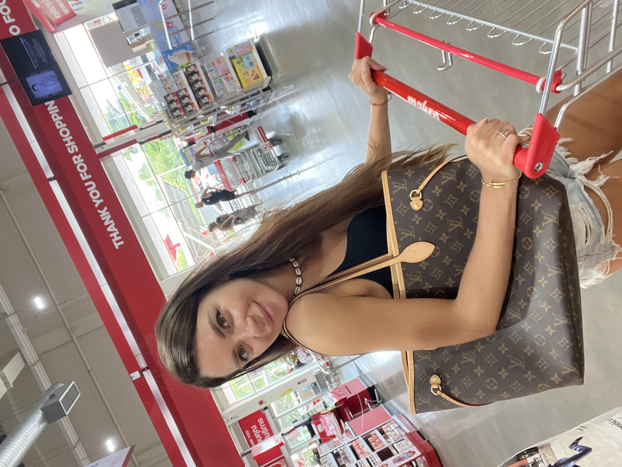 A woman with long brown hair and a white shell necklace poses for a selfie in a bright supermarket. She wears a black tank top and distressed denim shorts. In her right hand, she holds the red handle of a shopping cart, and her left arm cradles a large Louis Vuitton tote bag. Behind her, shelves stocked with various products are visible, along with other shoppers and shopping carts in the aisles. A large red sign overhead reads "THANK YOU FOR SHOPPING." The flooring appears to be a light-colored, polished concrete. The overall environment suggests a retail setting in Baan Hua Thanon, Thailand.