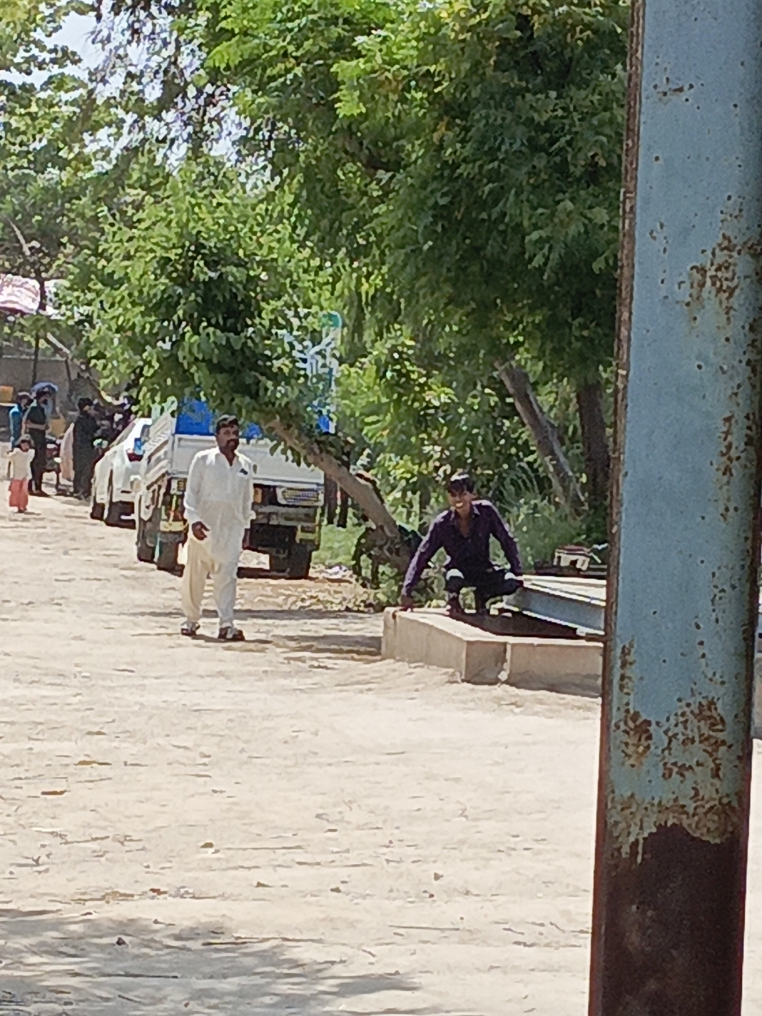 A bright, sunlit outdoor scene, likely a village or semi-rural area in Karamsar, Pakistan. The foreground features an unpaved, light-colored dirt road. On the far right, a tall, heavily rusted metal pole, predominantly light blue with reddish-brown corrosion, vertically frames the image.
In the mid-ground, a man in a white shalwar kameez and dark sandals walks towards the viewer on the dirt road. He has a beard. Behind him and to the left, a small child in a light top and pink lower garment walks away, and several other people are partially visible near parked vehicles. Two white pickup trucks or small cargo vehicles are in the background, partly obscured by people and dense green trees.
On the right side of the mid-ground, a man wearing a dark purple or maroon long-sleeved shirt and dark pants is crouching on a raised concrete structure. He looks towards the left with a slight smile. Beside him on the concrete are several large, light-colored metal beams or construction materials.
The background is lush with numerous green trees, indicating a verdant environment. The bright, direct light suggests a clear, sunny day, likely midday or early afternoon. No discernible text is visible in the image. The scene captures everyday activity on a sunny day in a natural, unpaved setting.