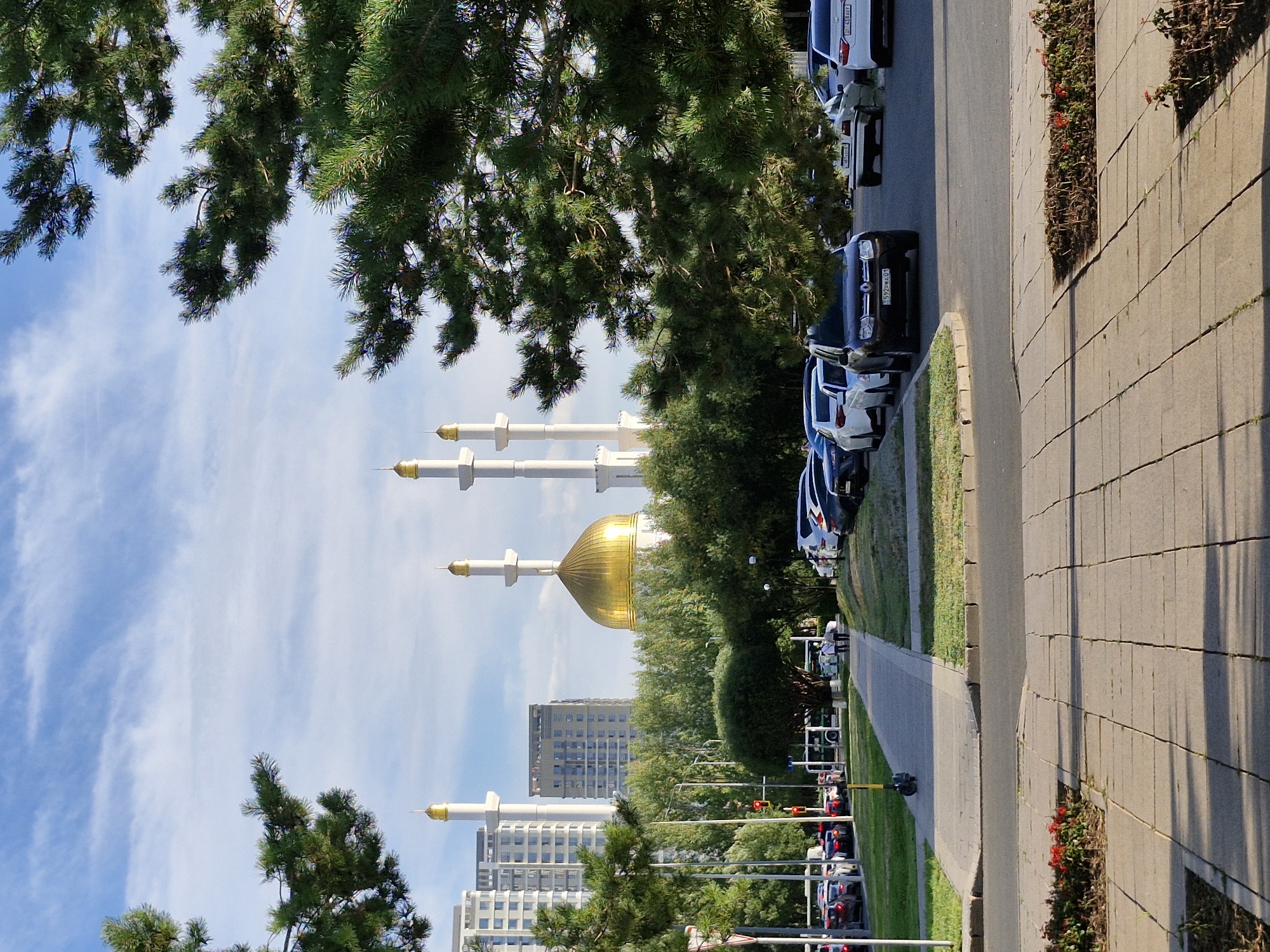 This is a vertical, eye-level, medium shot of a mosque in Astana, Kazakhstan, partially obscured by the branches of a coniferous tree in the foreground. The mosque, with its prominent gold dome and four white minarets topped with gold spires, is centrally located behind a row of trees and several cars parked on the right side of the frame.
To the left of the mosque, a cluster of modern, tall residential buildings with visible windows are situated. A street with a sidewalk and parked cars lines the foreground, and a paved walkway extends from the foreground sidewalk towards the mosque. Two figures, appearing to be adults walking side-by-side, are visible on this walkway in the middle ground.
The sky above is a pale blue, with scattered wispy white clouds. The overall lighting suggests it is daytime, with sunlight casting shadows on the paved areas in the foreground. The scene depicts an urban environment with a focus on the mosque as a significant architectural landmark.