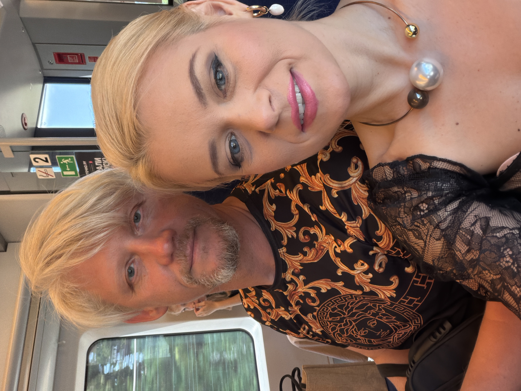 The image shows a close-up, selfie-style shot of two people, a man and a woman, in what appears to be a train carriage. The man, in the foreground, has blonde hair and a light beard, and is looking towards the camera. He is wearing a black shirt with a gold baroque-style print. The woman, partially behind and above him, has blonde hair and is also looking at the camera with a bright smile. She is wearing makeup, including pink lipstick and eyeliner, and a distinctive necklace with large pearl-like beads. She also has on a black lace garment.
The setting appears to be daytime, as indicated by the light coming through the train window, which shows greenery outside, suggesting the train is in motion. Interior train features, such as a window, seats, and signage are visible. One sign has Cyrillic text and appears to indicate an emergency exit. The overall mood seems casual and perhaps even celebratory, given the woman's makeup and smile, and the couple's close proximity.
Visible text includes what appears to be "ТОЧНО ВУ" and other Cyrillic characters on a sign. The overall impression is a candid moment captured during a train journey in Sochi, Russia.