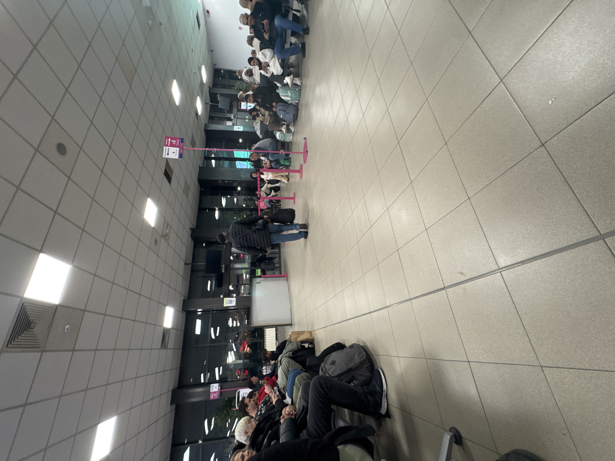 The image shows an airport waiting area, likely in Sofia, Bulgaria based on the context. Passengers are seated in rows of chairs, with luggage placed nearby. Some people are standing in a line, possibly waiting to board or for security. The environment is sterile and modern, with tiled floors and fluorescent lighting. The scene suggests travel and anticipation.