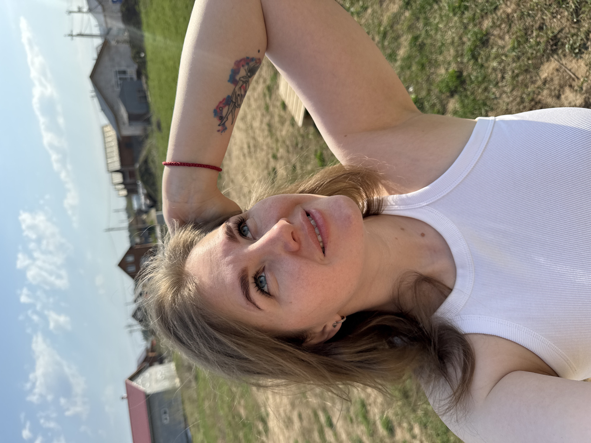 A woman is outdoors in Ферма, Russia. She has fair skin, blue eyes, and light brown hair. She is wearing a white ribbed tank top. Her left arm is raised, with a red string bracelet visible on her wrist and a colorful tattoo on her forearm. The background includes grass and buildings, with a partly cloudy sky overhead.