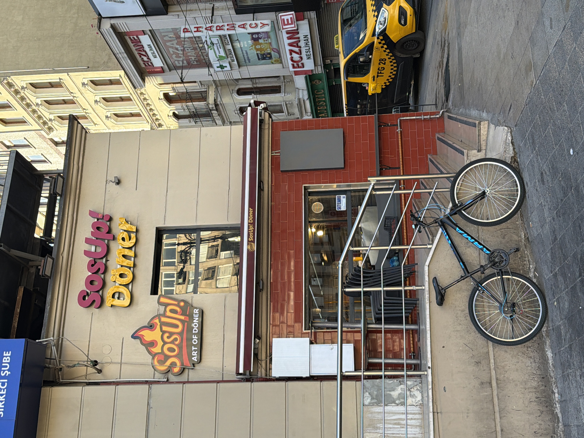 The image captures a street scene in Istanbul, Turkey, with a focus on a restaurant named "SosUp! Döner" and its immediate surroundings. The restaurant's facade features prominent signage in pink and yellow, with the words "SosUp! Döner" and "ART OF DÖNER." A window reflects buildings across the street. Adjacent to the restaurant, several buildings are visible, including those with signage for "ECZANE" (pharmacy) and "PHARMACY." A yellow taxi with "TFG 28" visible on its side is parked on the street. In the foreground, a black bicycle with blue accents is leaning against a concrete wall, partially obscuring a set of stairs leading up to the restaurant entrance. The scene appears to be on a street with brick and concrete pavement. The overall impression is a typical urban streetscape in Istanbul, showcasing commercial establishments and everyday street life.