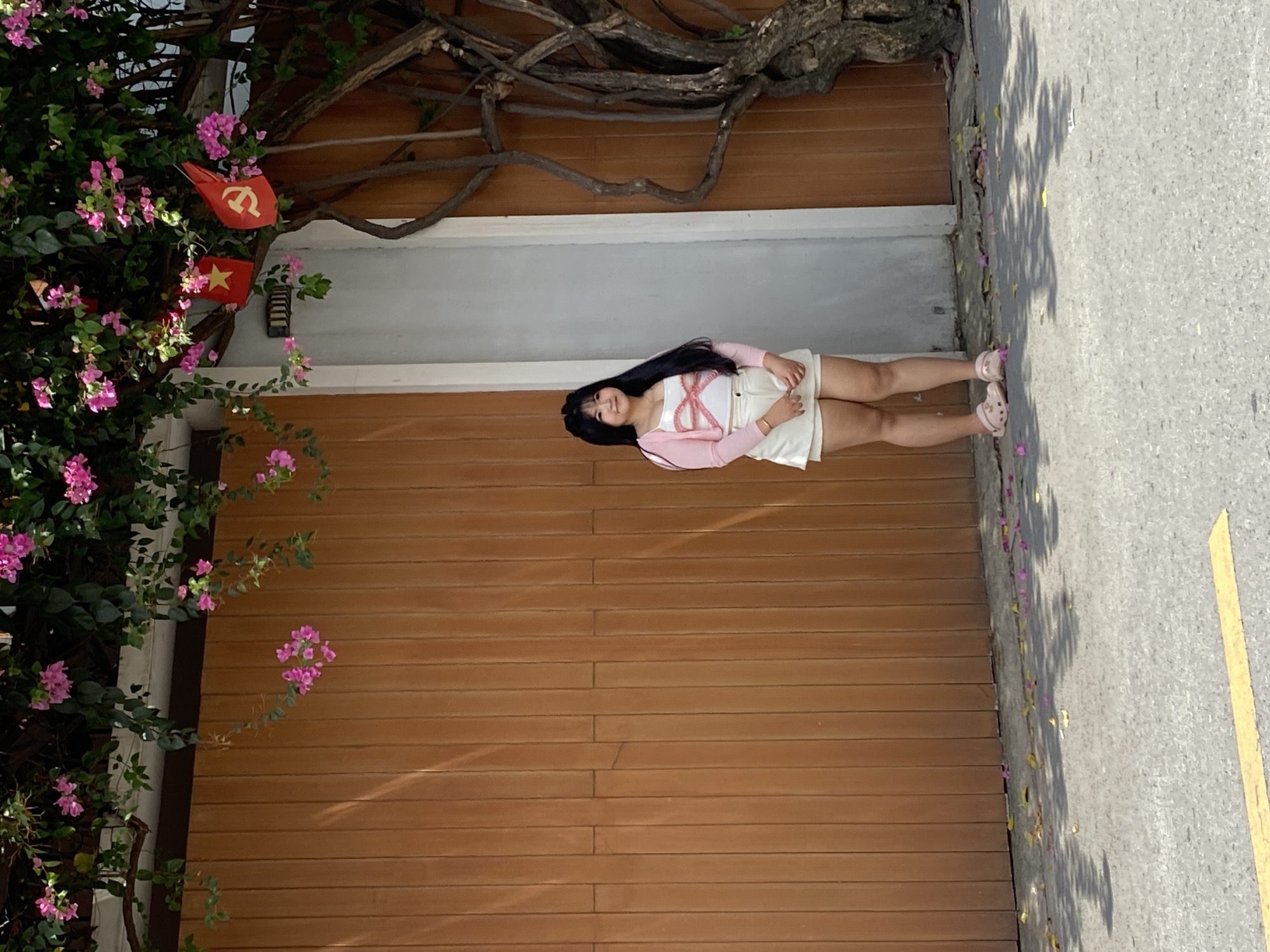 A young woman with long, dark hair is lying on a wooden deck in front of a building in Đồng Nai, Vietnam. She is wearing a white top, a pink cardigan, and white shorts. She is facing the camera and smiling slightly. To the left of the deck is a bush with pink flowers. Above the deck is a tree branch with a few leaves. In the background, there is a gray concrete street with a yellow line. Vietnamese flags are hanging on the building.