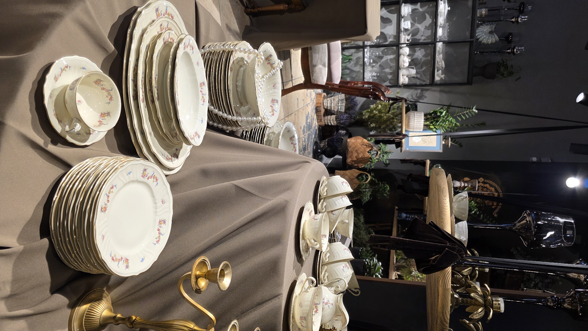This is a medium shot, eye-level, indoor photograph of a display of fine china and decorative items. The scene is set against a dark gray wall and on tables covered with brown fabric. In the foreground, a stack of white dinner plates with floral patterns and gold trim is prominently displayed. To the left, a white teacup and saucer with a similar floral design sits on the table. Further back, a large serving bowl is stacked on a set of plates, adorned with a strand of pearls. Several teacups and saucers are arranged on a round table covered in the same brown fabric, some containing beverages. A white pitcher with a floral design is also visible. A gold-colored candelabra with two candle holders stands on the right side of the frame. In the background, a black metal shelf unit displays various glassware and decorative objects. A wooden chair with upholstered seating is positioned behind the first table. Hanging from the ceiling are black straps supporting wooden shelves with potted plants. The lighting is dim, with spotlights illuminating parts of the display. There are no people visible in the image, and no specific location in Riga, Latvia can be identified.