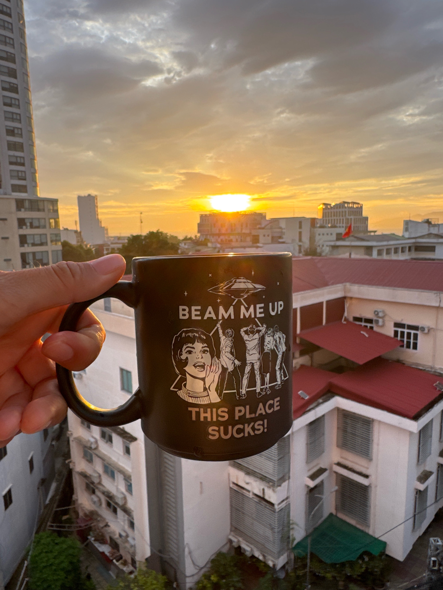 An individual's hand holds a dark mug with white text and graphics, "BEAM ME UP THIS PLACE SUCKS!", in front of a city skyline at sunset. The mug depicts a flying saucer and silhouetted figures, one of which appears to be a stylized drawing of a woman. The background features buildings of varying heights, including a tall, modern high-rise on the left, and a complex of white buildings with red roofs. Trees and clouds are also visible, with the sun setting on the horizon, casting a warm orange glow. The scene is consistent with a view from a high vantage point within Nha Trang, Vietnam, given the architecture and tropical vegetation visible.