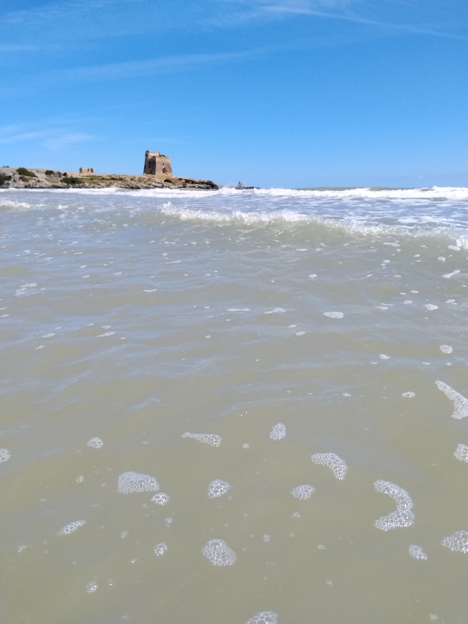 The image depicts a serene coastal scene with a stone tower on a rocky outcrop overlooking the sea. The tower, appearing ancient and weathered, stands prominently against a clear blue sky. The foreground is dominated by the gentle lapping of waves, with some foam and bubbles visible on the surface of the pale, somewhat murky water. The horizon is marked by the meeting of the sea and the sky, with a faint hint of a distant landmass or structure on the left side of the image. There are no people or animals visible in the scene. This scene is reminiscent of the coastline near Peschici, Italy, known for its picturesque towers and rugged shores.