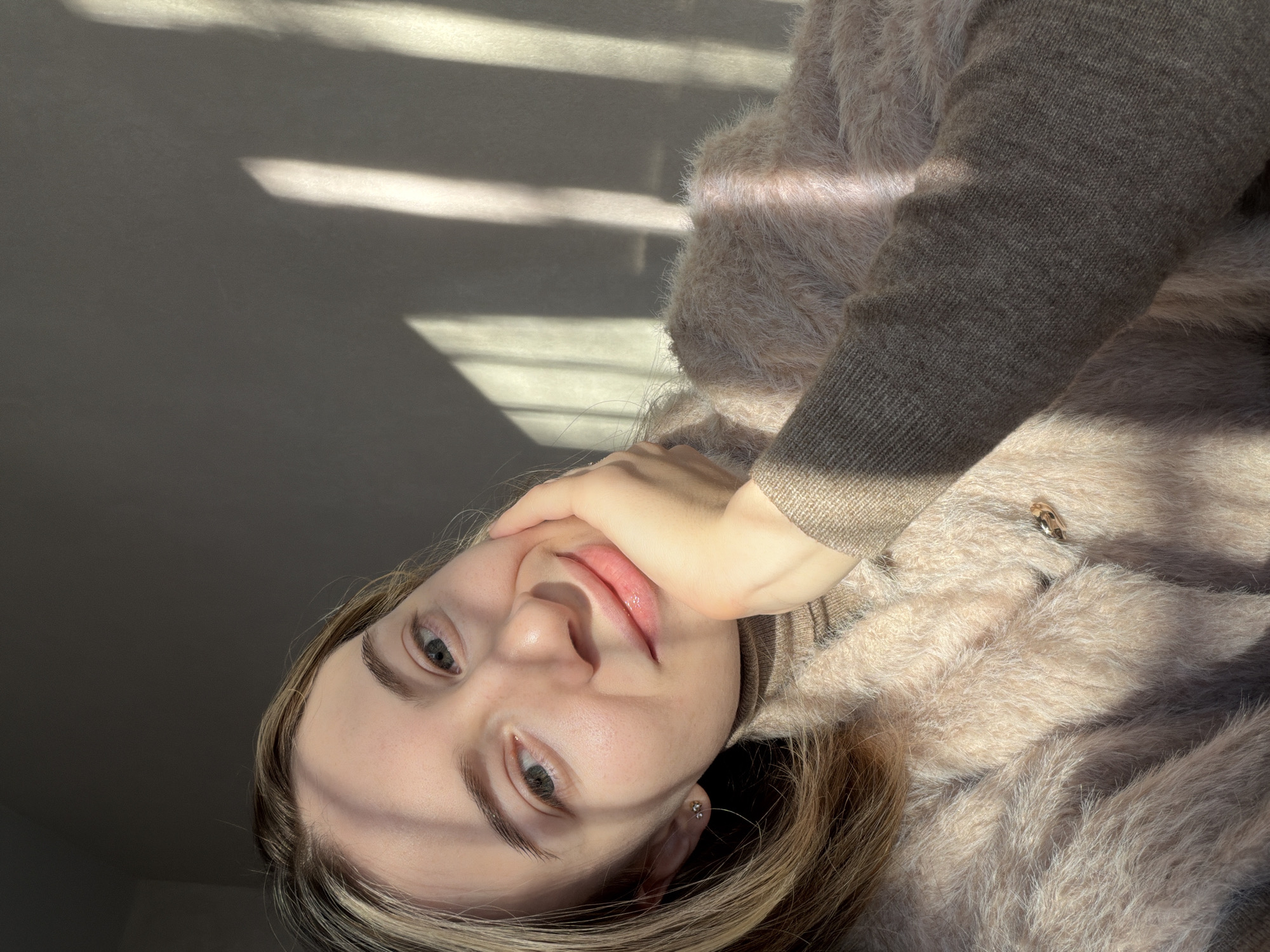 A woman is posing for a selfie, her face angled upwards towards the camera. She has light skin, light eyes, and light brown hair with blonde highlights. Her right hand is touching her face, near her cheek. She is wearing a brown turtleneck sweater and a fluffy, beige cardigan with a visible button closure. The background is a plain gray wall, with streaks of sunlight. The location of the photo is impossible to determine based on visual information alone.
