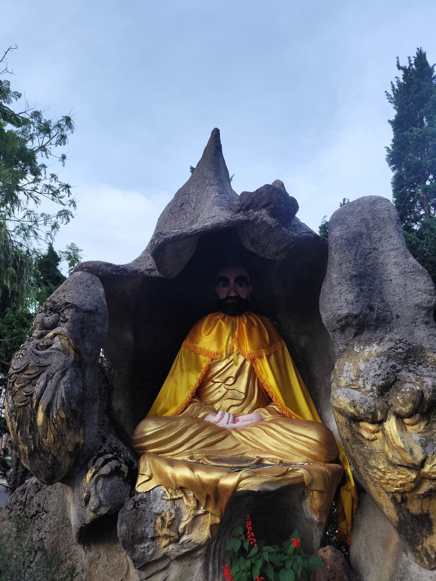 A ground-level shot showcases a golden statue of a seated figure in a meditative pose. The figure is clothed in a golden robe with yellow trim. The statue is housed within a rocky grotto, with gray stone formations surrounding it. The formation above the statue's head has a pointed peak. To the left of the formation, there's a rock sculpted into a lion's head. To the right, a simple gray stone formation. At the statue's base, there are small red flowers among green foliage. The background shows trees and a cloudy sky. The location is Cam Ly - Đà Lạt, Vietnam.