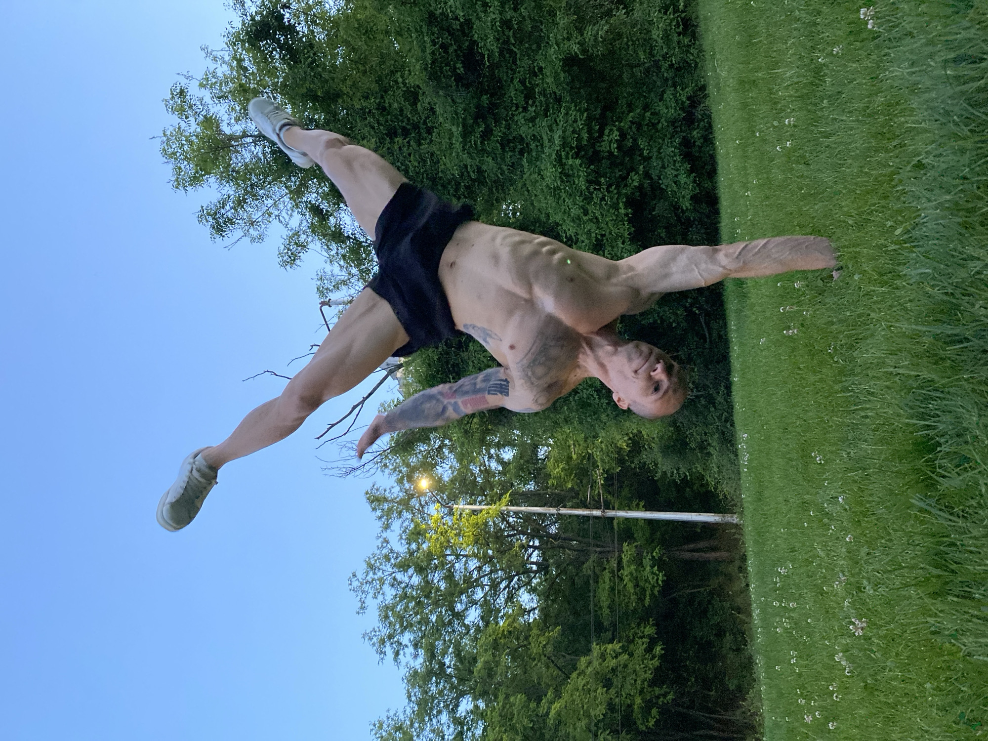 An adult male is performing a handstand split in an outdoor location, likely a park in Pleven, Bulgaria. He is wearing black shorts and light-colored sneakers. His body is toned, and he has tattoos on his arm and chest. One hand is planted on a green lawn with small white flowers, and the other hand is extended to the side. His legs are split, one pointing upwards towards a light blue sky partially obscured by tree branches, and the other pointing towards more green trees. A lamppost is visible in the background.