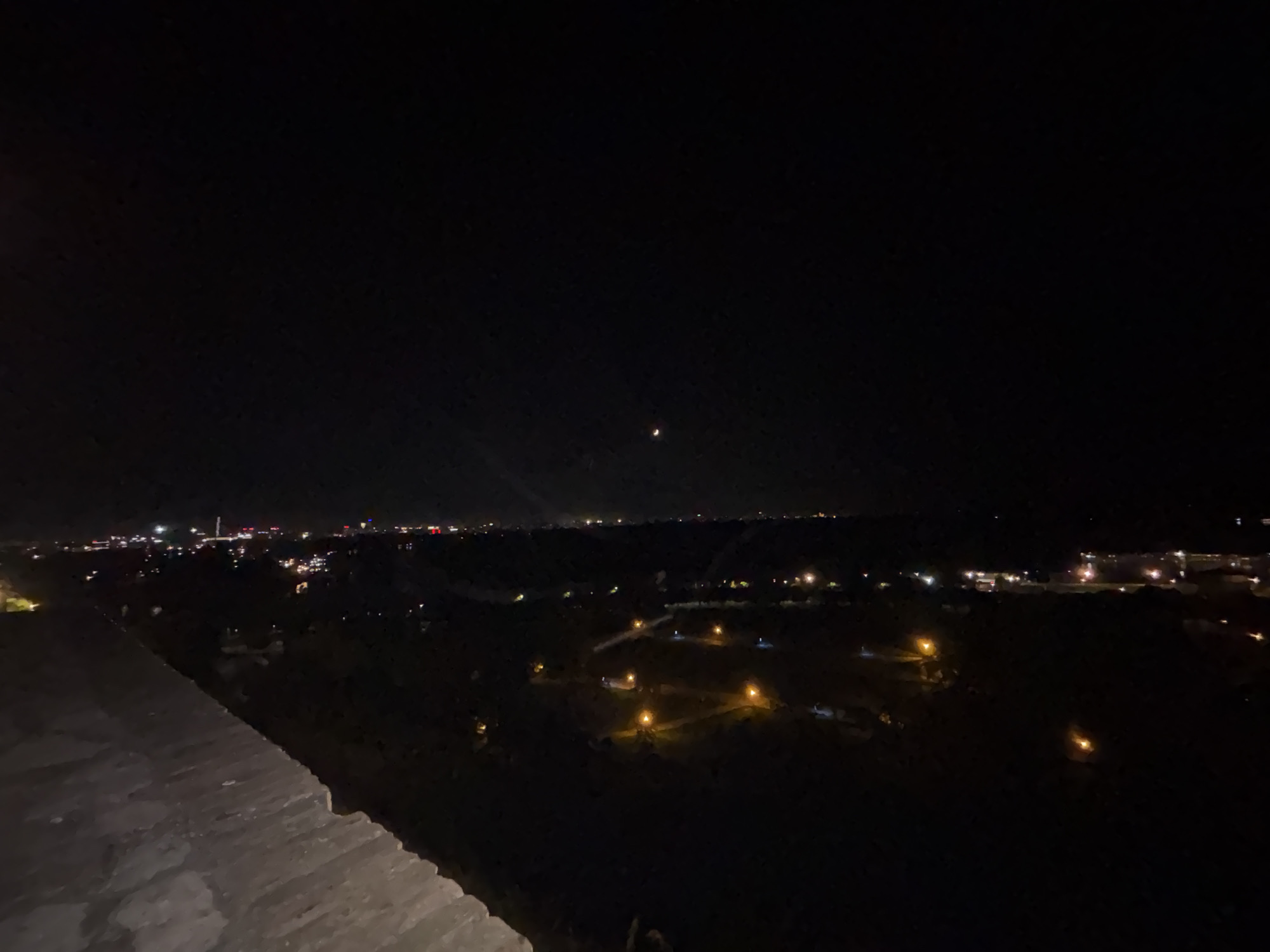 Night scene of Belgrade, Serbia. From an elevated position, a dark cityscape is visible, interspersed with scattered lights indicating residential and commercial areas. In the distance, city lights form a faint horizon. Close by, a stone structure is partially illuminated, likely a wall or part of a building. The night sky is dark, with a crescent moon visible. The environment is quiet.