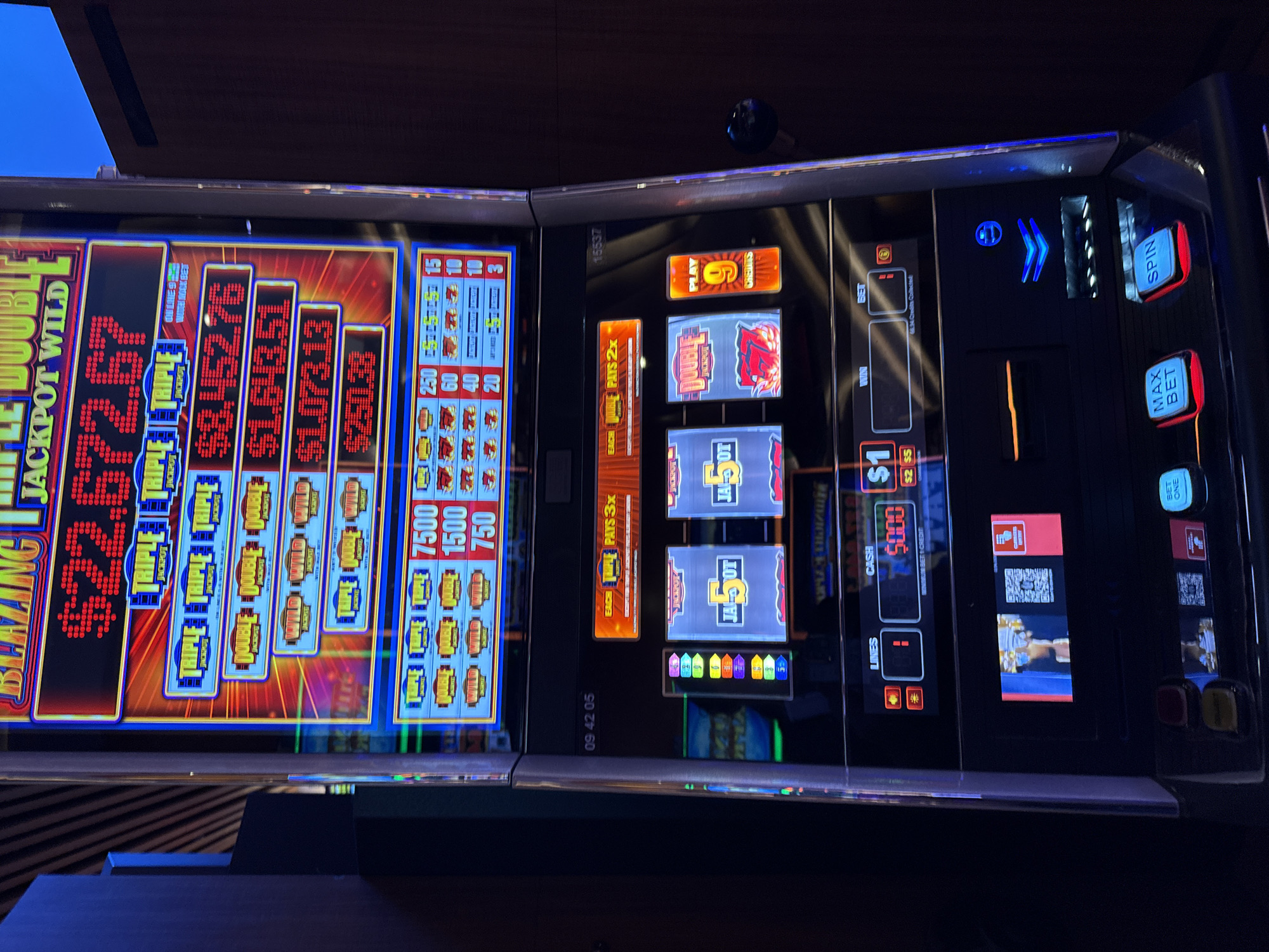 The image shows a close-up of two slot machines in what is likely a casino. The left slot machine displays "BLAZING JACKPOT WILD" and various jackpot amounts, including $22,672.67, $1,643.51, $1,073.13, and $250.38. The right slot machine shows the reel window with "5 J-A O-T" visible. A display shows $5,000 as the current cash amount. Control buttons such as "SPIN", "MAX BET," "BET ONE," and a pay table section are visible. The timestamp "09 42 05" is present. The image was taken in the city of Hollywood Seminole Reservation, United States of America.