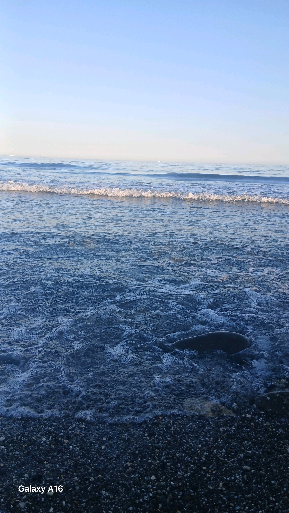 The image shows a serene beach scene at what appears to be dawn or dusk. The sky is a pale blue, fading to a lighter hue near the horizon. Gentle waves with white foam crests are rolling in towards a pebble beach. In the foreground, a large, dark, smooth stone is partially submerged in the shallow water, with smaller pebbles making up the shoreline. The water is a deep blue, with a foamy texture from the breaking waves. In the distance, two small sailboats can be seen on the calm sea. The overall impression is one of tranquility and natural beauty. It is not possible to determine the specific location in Marbella from the image alone.