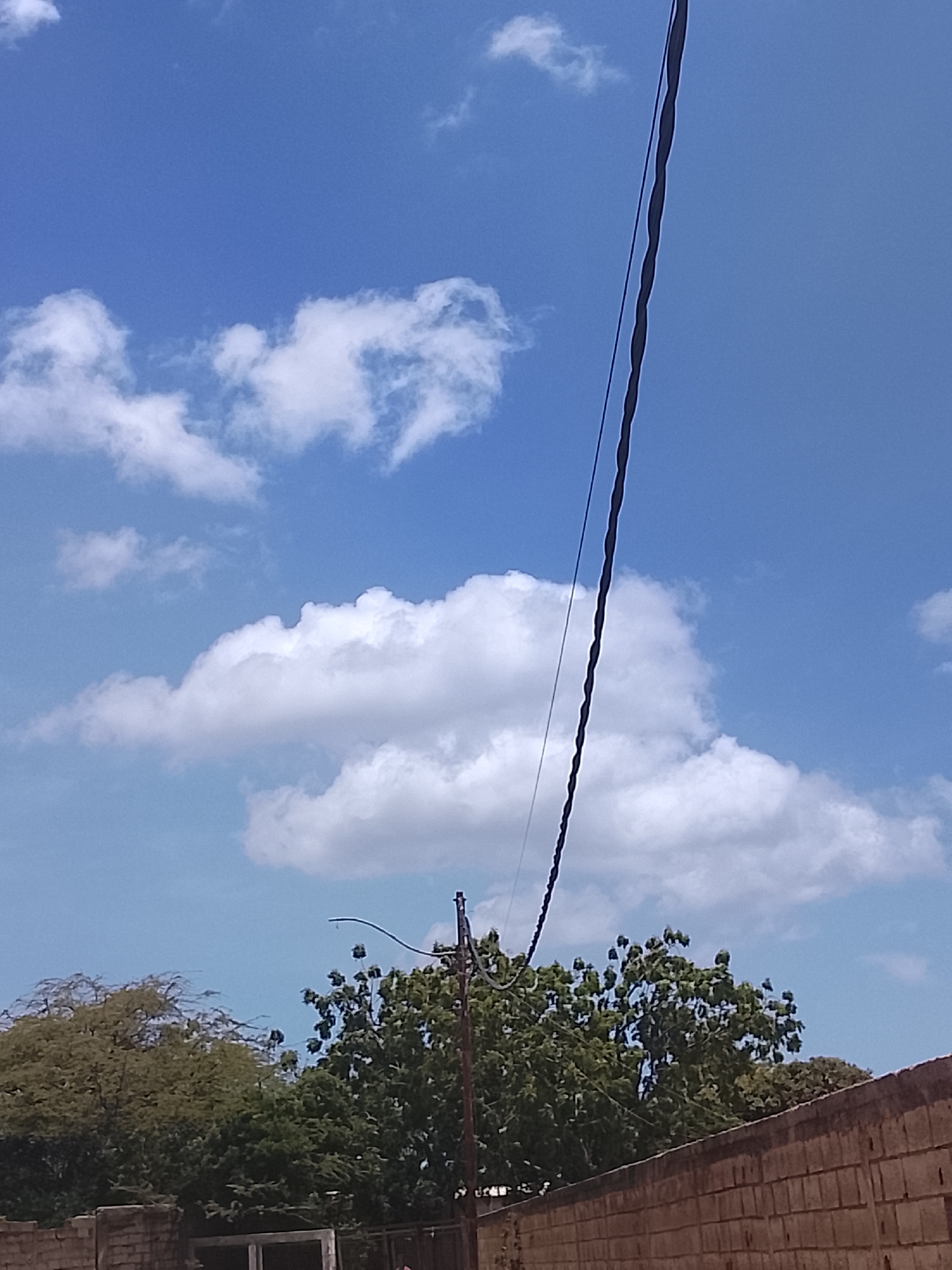 The image depicts a partly cloudy sky with visible power lines running diagonally across the frame. Below the sky, there are trees, with a brick wall visible at the bottom of the image. This could be in the city of Altagracia, Venezuela, but there is no definite proof.