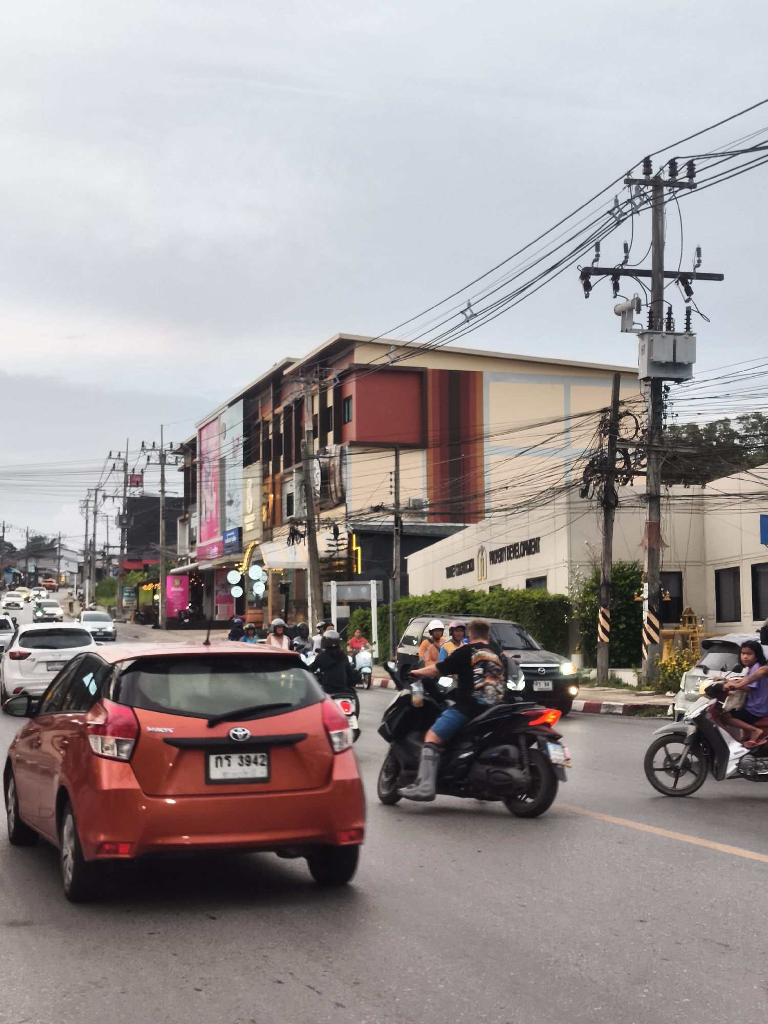 The image depicts a street scene in the Ko Samui District of Thailand, likely during daytime, with overcast skies. The foreground is dominated by the rear of an orange hatchback car. Traffic is present, with several other cars and numerous motorcycles on the asphalt road. On the right side of the road, a white scooter carries a passenger wearing a purple top. On the left, a black scooter with two occupants is in motion. Behind the traffic, there are multi-story buildings lining the street, featuring a mix of commercial and residential designs with balconies and signage. Electrical poles with numerous wires are prominently visible throughout the scene, indicating urban infrastructure. The overall impression is of a busy commercial street in a tropical climate.