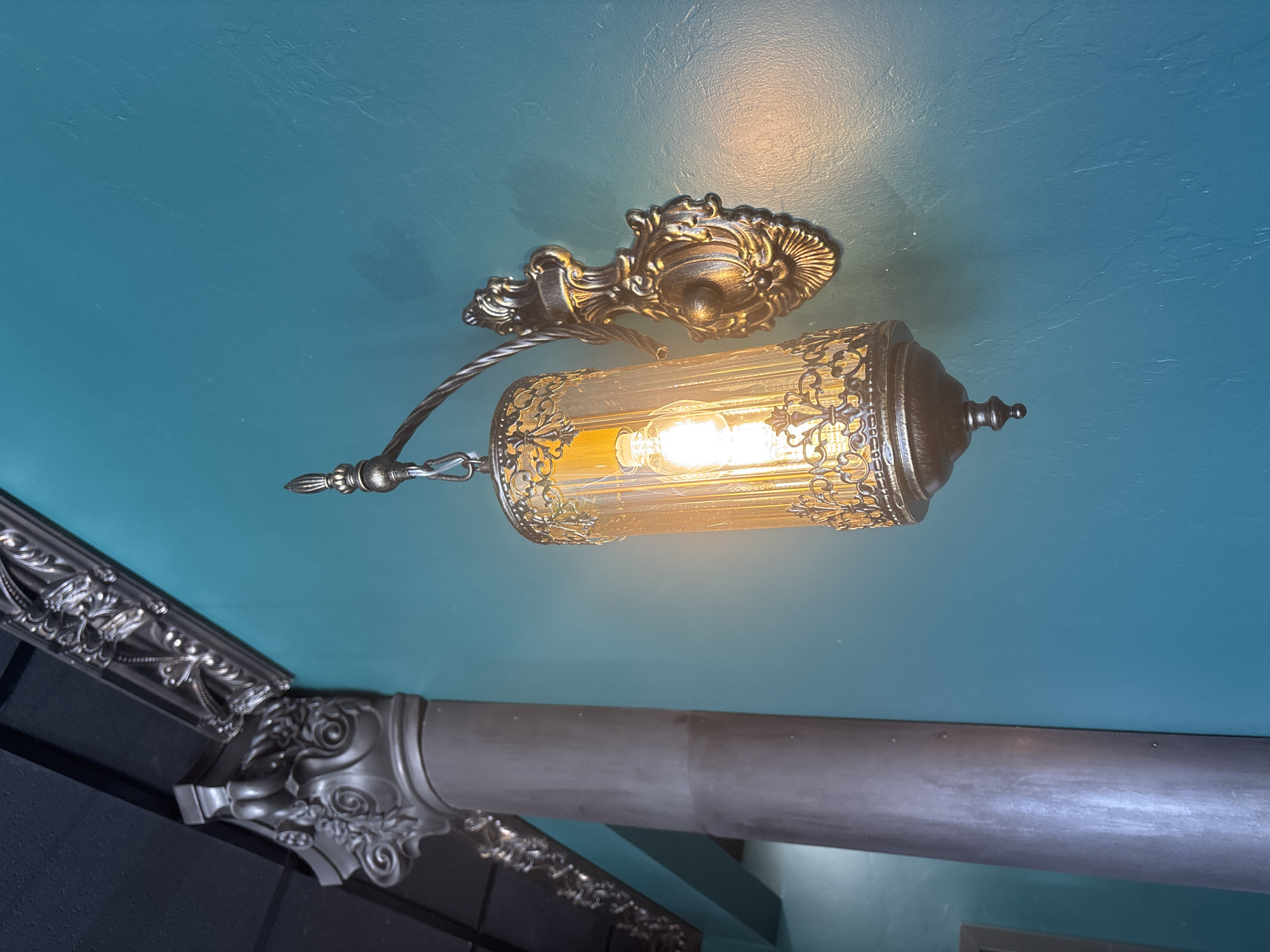 An ornate, vintage-style wall sconce with a clear, ribbed glass shade and intricate metalwork is mounted on a textured, teal-colored wall. The sconce has an exposed Edison bulb that emits a warm, yellow light. To the left of the sconce, a decorative, fluted column with metallic trim and intricate carvings is visible, partially obscuring a dark, possibly wooden ceiling beam with decorative molding. The lighting suggests an indoor setting, likely a restaurant or decorative interior space. The image does not provide any clues about its location in Tucson, United States of America.