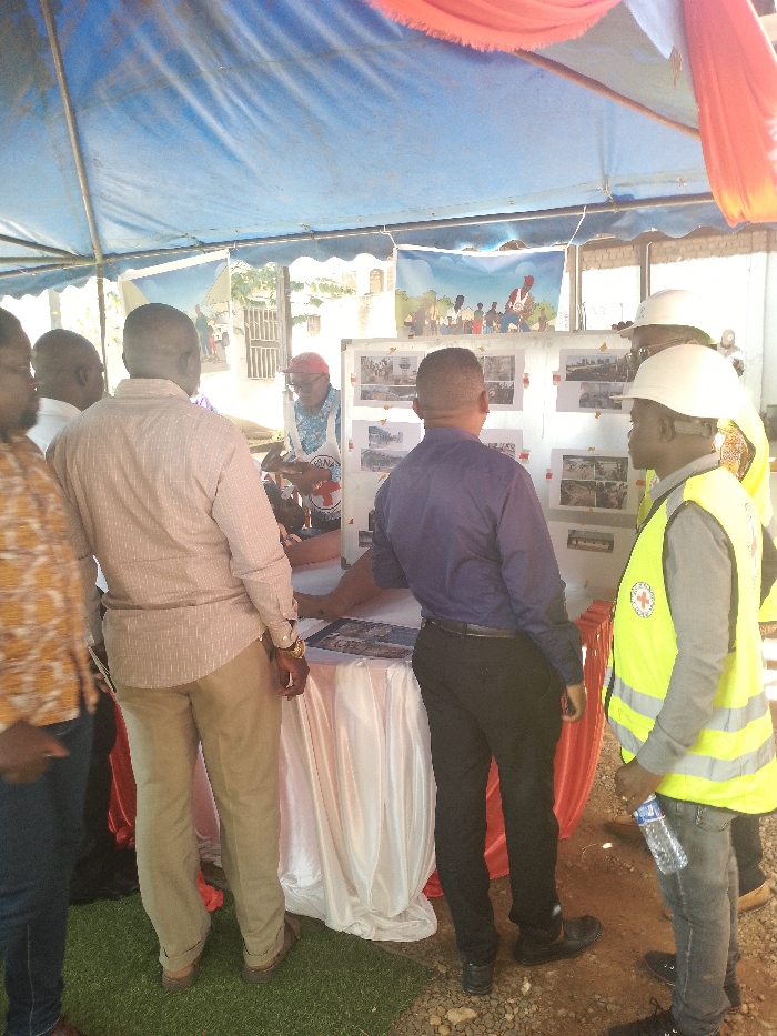 A group of six men stand around a table covered with a white tablecloth. One man, wearing a white shirt and a red vest with a red cross printed on it, stands to the left of a white board displaying photos, seemingly pointing at one. Three men stand to the right of the board, one wearing a bright yellow vest, the other in a blue shirt, and the last in a white hard hat with a white vest. Another man with a red vest is partially visible behind the board. The men are gathered under a tent, and a red tablecloth is visible to the right. The tent is open, and a small patch of green grass is visible behind the table. There is an outdoor setting behind the tent. The scene likely depicts a meeting or event related to the International Committee of the Red Cross (ICRC), as evidenced by the red cross logo worn by some participants. The location is Bunia, Democratic Republic of the Congo, specifically at coordinates latitude: 1.556, longitude: 30.244.