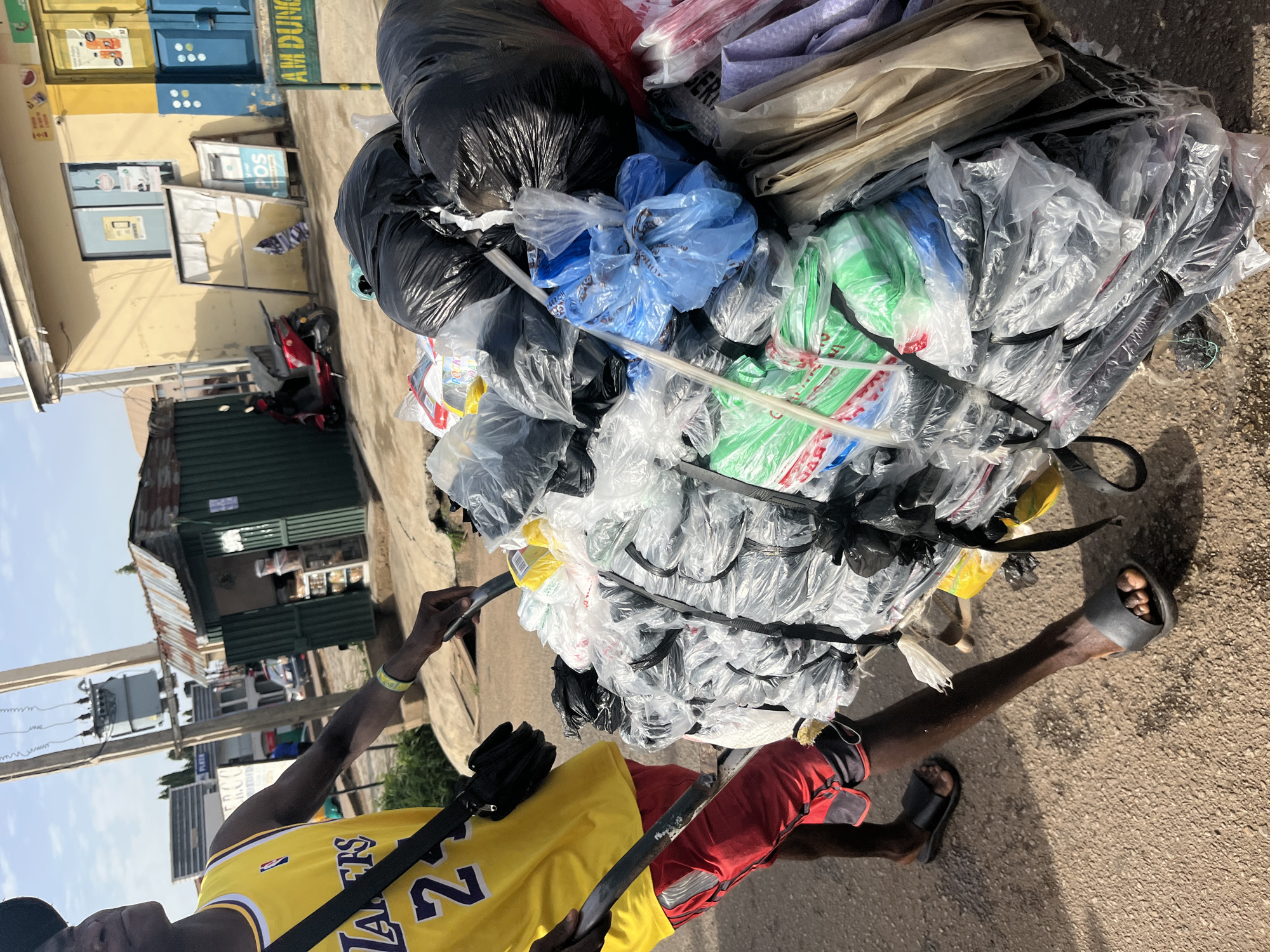 A young man in a yellow basketball jersey with the number 23 and red shorts pushes a cart laden with black and clear plastic bags. He is walking on a paved street in Jos, Nigeria. The cart is full of various plastic bags, some transparent, some black, and some colored green and blue. In the background, there are buildings, a red motorcycle parked, and a street sign that reads "AM DUNG". The sky is partly cloudy. The man is wearing black sandals.
