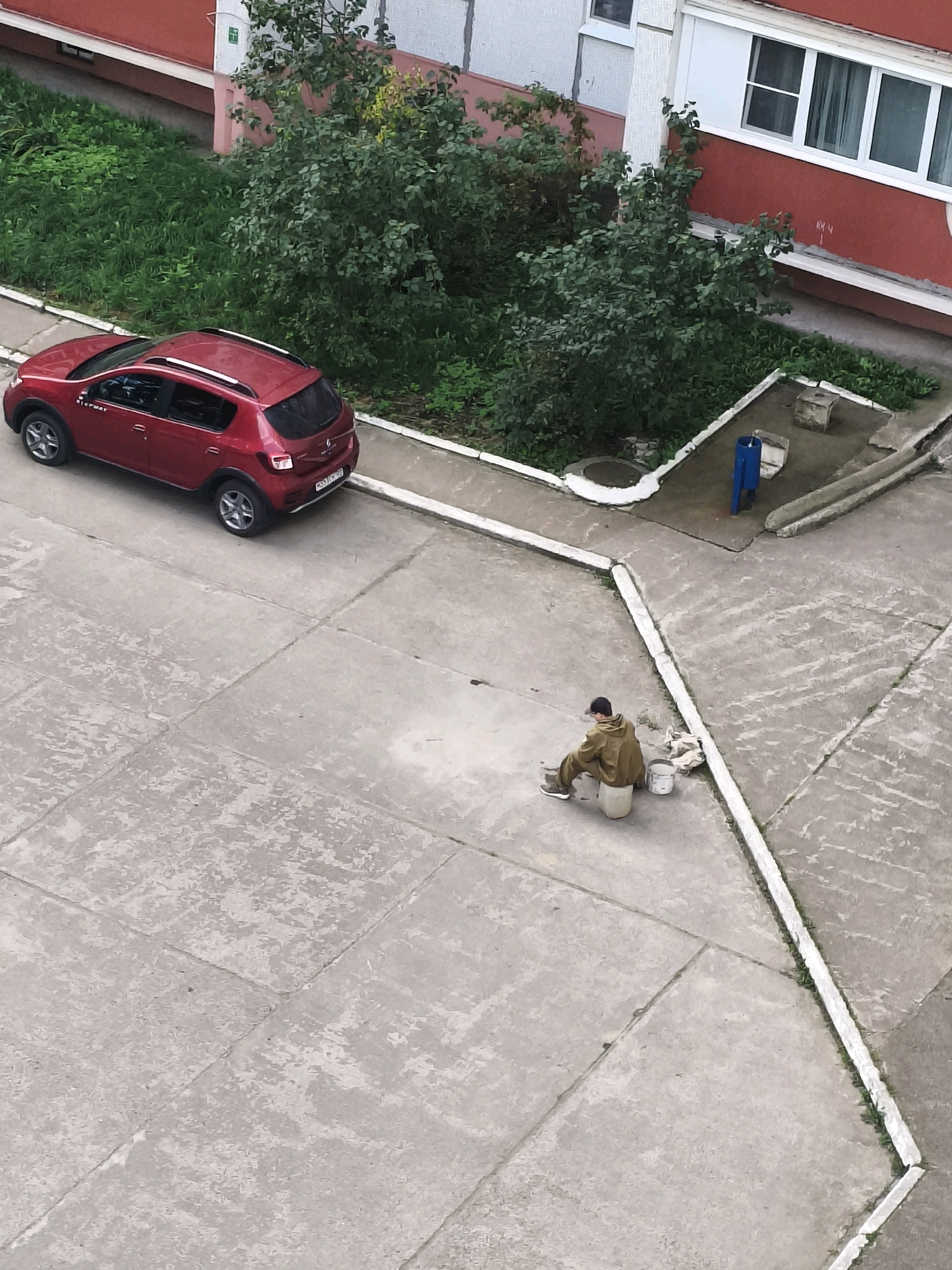 An aerial, high-angle, full shot shows a red Renault Sandero Stepway parked in a concrete courtyard in Chaikovsky, Russia. To the left of the car is a grassy area. To the right of the car is a building with a red facade and white window frames. A person wearing a tan jacket and a cap is sitting on a white bucket in the courtyard to the right of the car. Next to the person are two white buckets. The person appears to be holding a brush. In the background is a small blue structure with a water faucet.