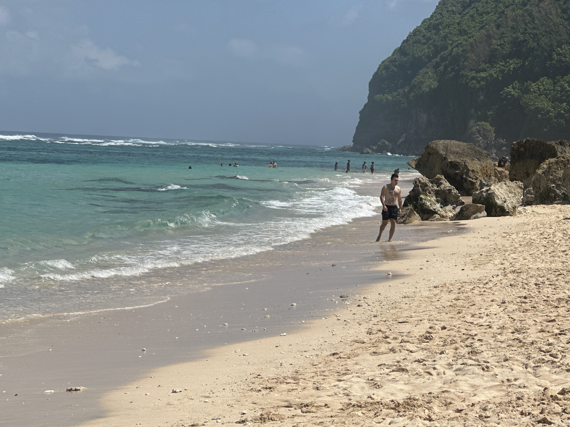 The image depicts a beach scene in Kuta Selatan, Indonesia. A fair-skinned man is walking along the shoreline. In the background, there is a group of people swimming or standing in the ocean. A rocky outcrop is seen to the right of the man. Lush green vegetation covers the cliffs forming the backdrop of the beach. The sky is overcast. The ocean has noticeable waves. The beach has a sandy shoreline.