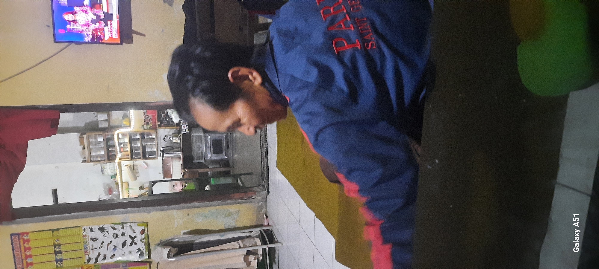 The image captures an indoor scene, likely a living room, in Ploso, Indonesia. A man is seated, facing away from the camera and towards a television screen displaying what appears to be a news program with a female anchor. The man is wearing a blue jacket with red lettering that reads "PARIS SAINT-GERMAIN". In the background, through a doorway, a kitchen area is visible, featuring shelves stocked with jars and containers. To the left of the man, a chart with various insects is displayed on the wall. The overall environment suggests a modest, lived-in domestic space. The Samsung Galaxy A51 watermark is visible in the bottom left corner.